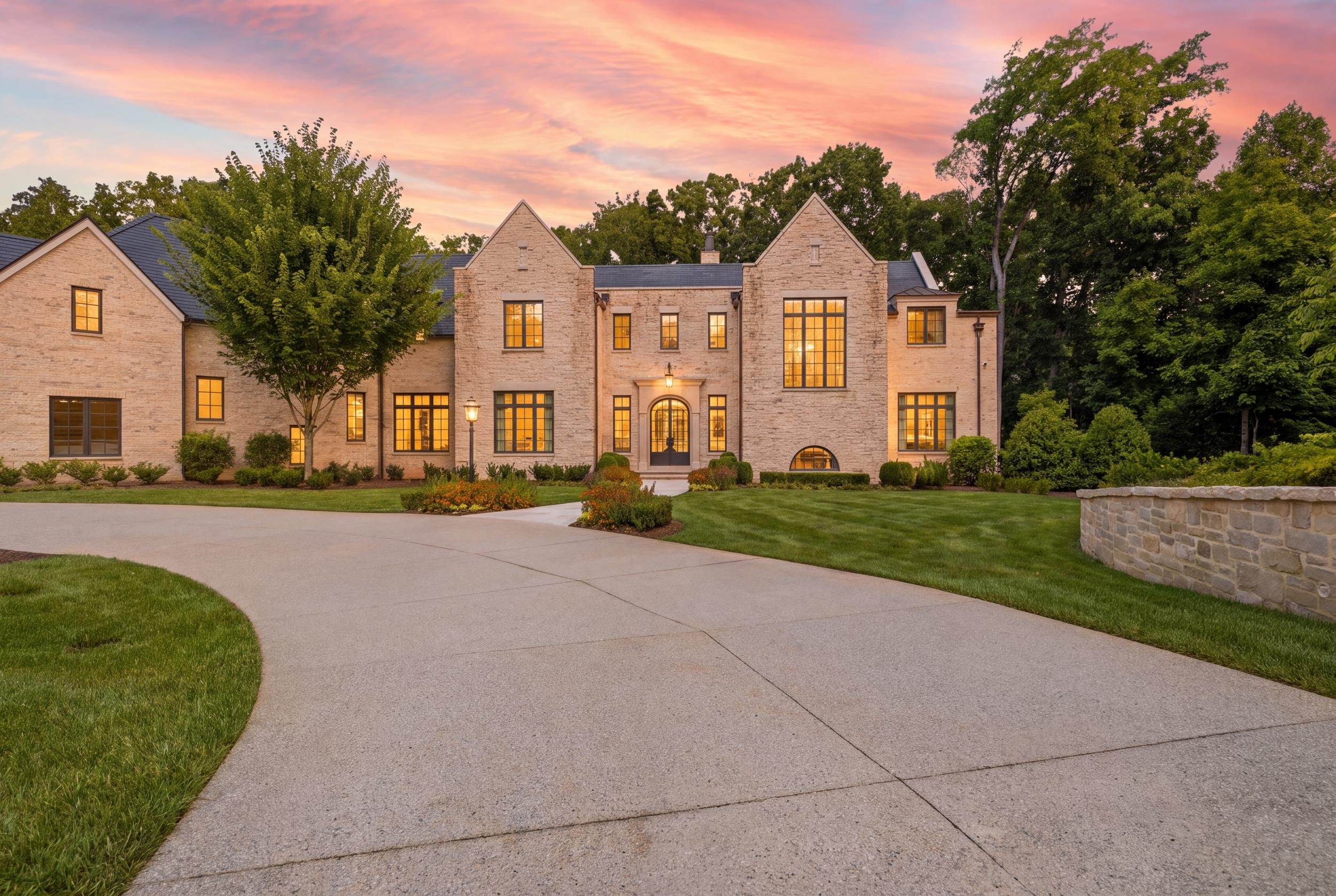 A large brick house with multiple peaked roofs, large windows with warm lighting, and a front door with an arched window above. The house is surrounded by a well-maintained lawn, trees, and landscaping. The sky has a colorful sunset with pink and ora