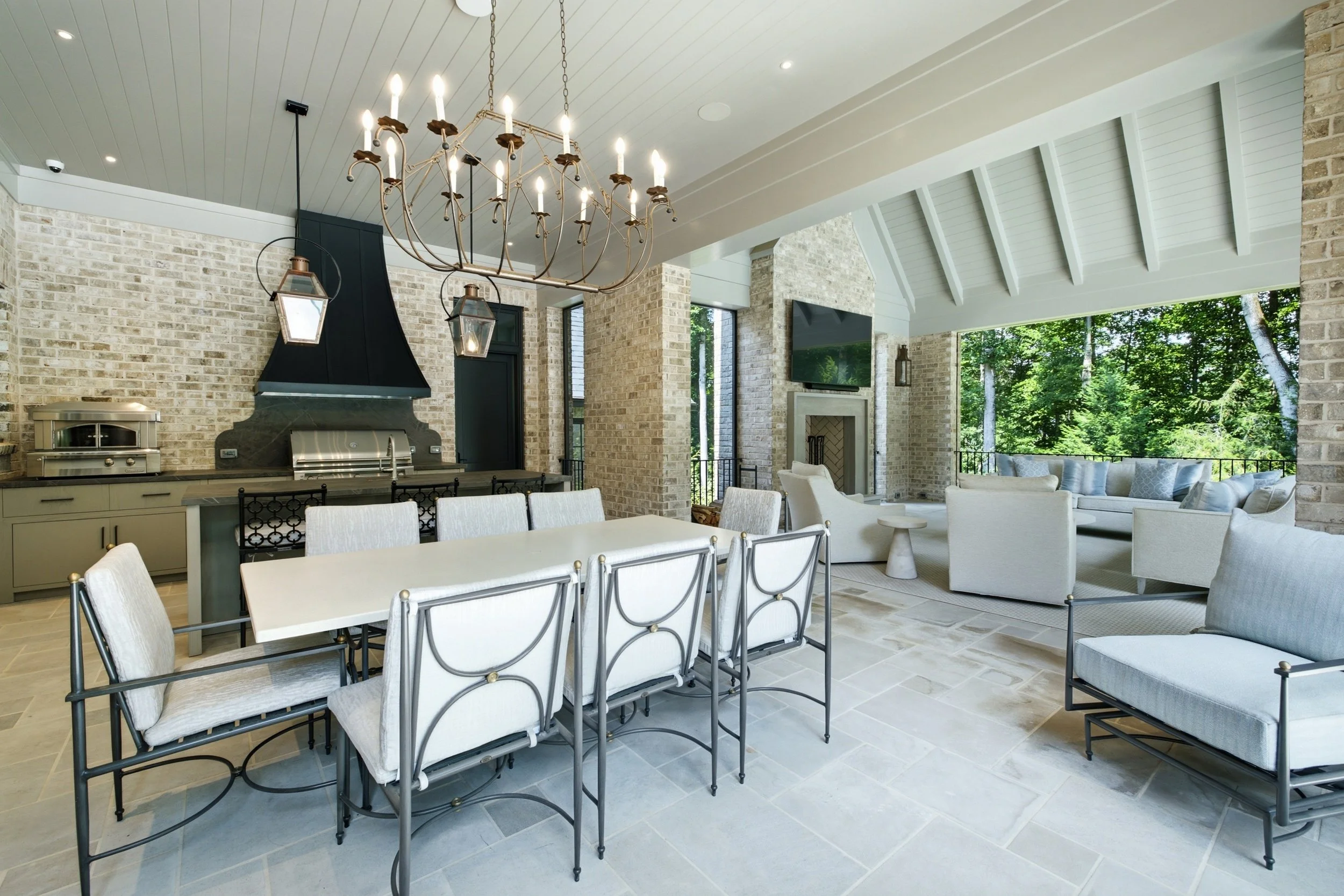 Interior of a spacious, bright living and dining area with a chandelier, brick walls, large windows showing green trees outside, white furniture, and a TV mounted on the wall.