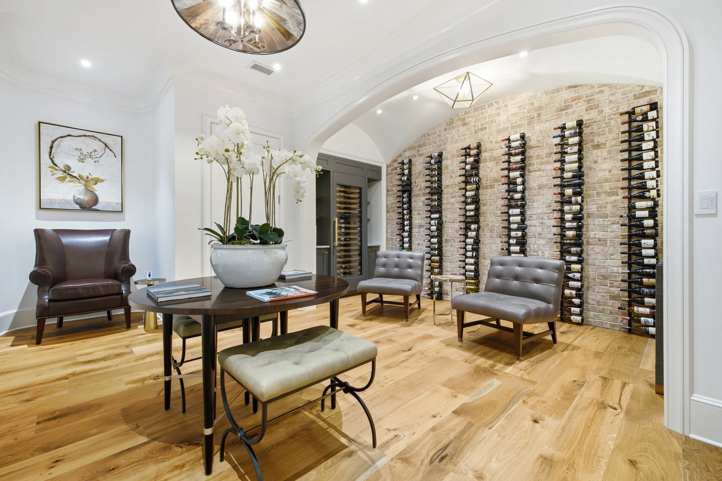 A cozy wine cellar with a brick wall, multiple horizontal racks filled with wine bottles, and a seating area with plush chairs. A round wooden table with a white orchid plant in a pot and magazines is in the center. There are framed art pieces on the