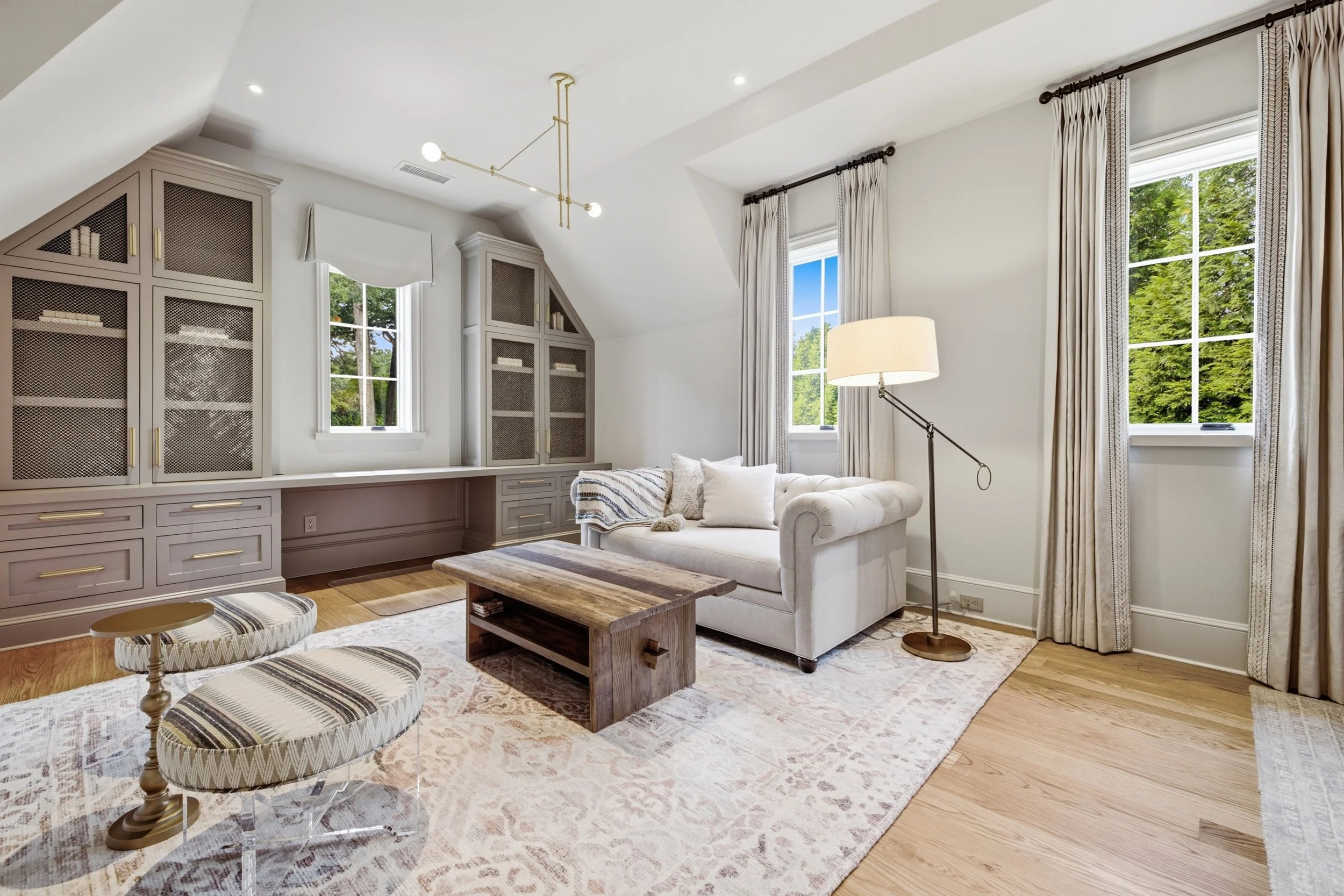 Light filled living room with white walls, large windows with cream curtains, a white tufted sofa with pillows, a wooden coffee table, patterned area rug, floor lamp, built-in white cabinets with gold handles, a modern chandelier, and hardwood floori