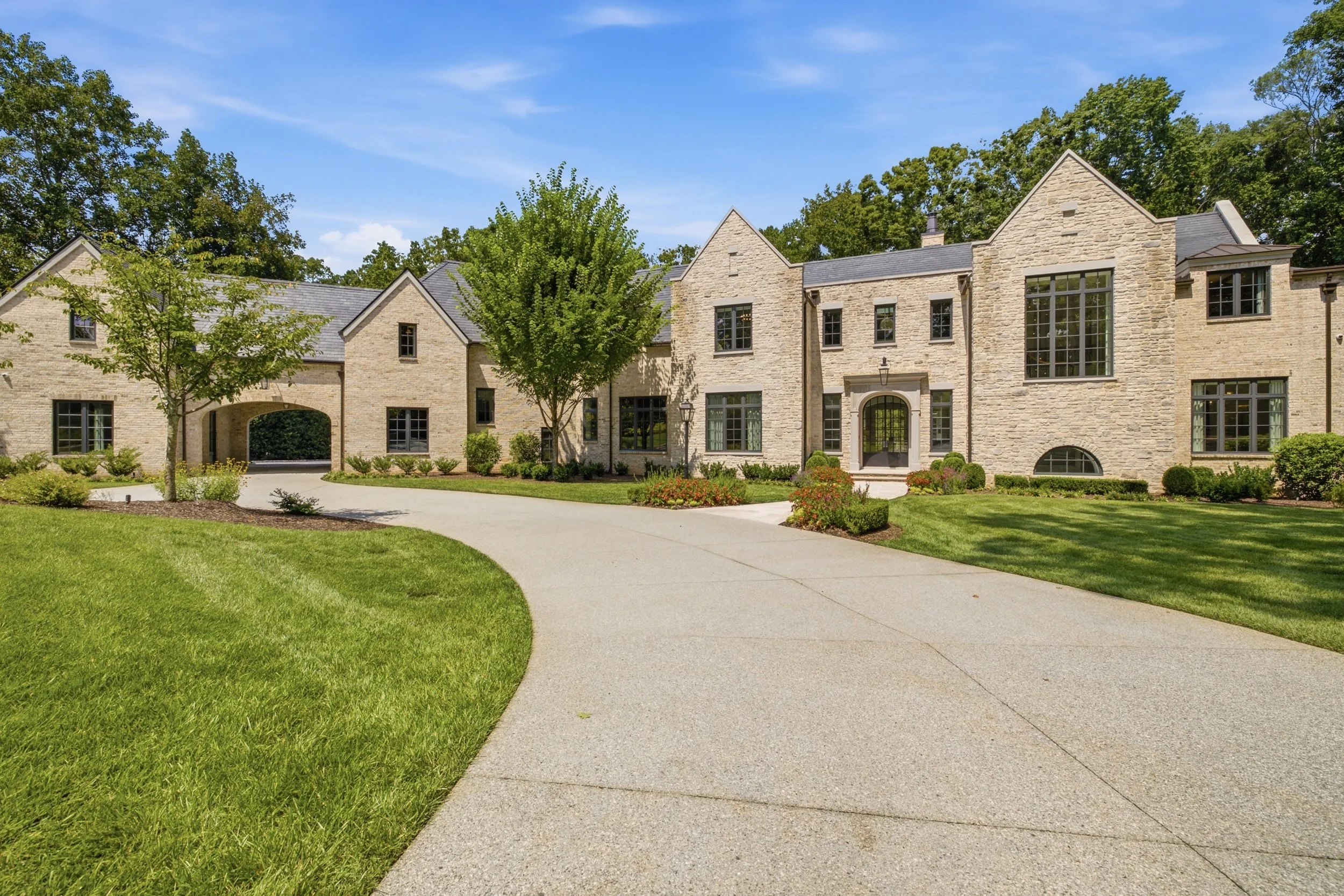 Large beige brick house with dark gray roof, multiple windows, and a curved driveway, surrounded by green lawn and trees under a blue sky.
