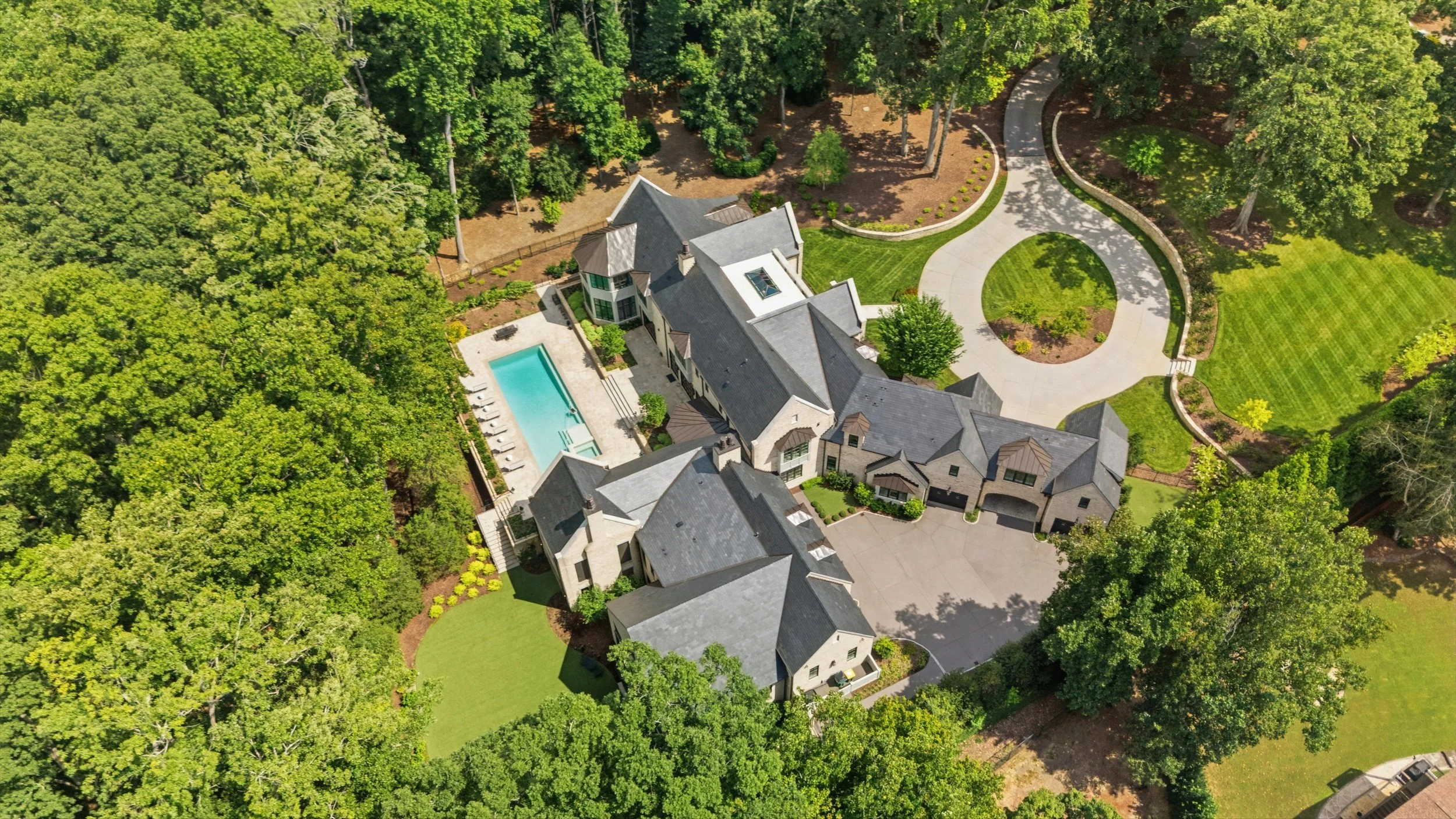Aerial view of a large house with a swimming pool, surrounded by lush green trees and landscaped yard, with a circular driveway.
