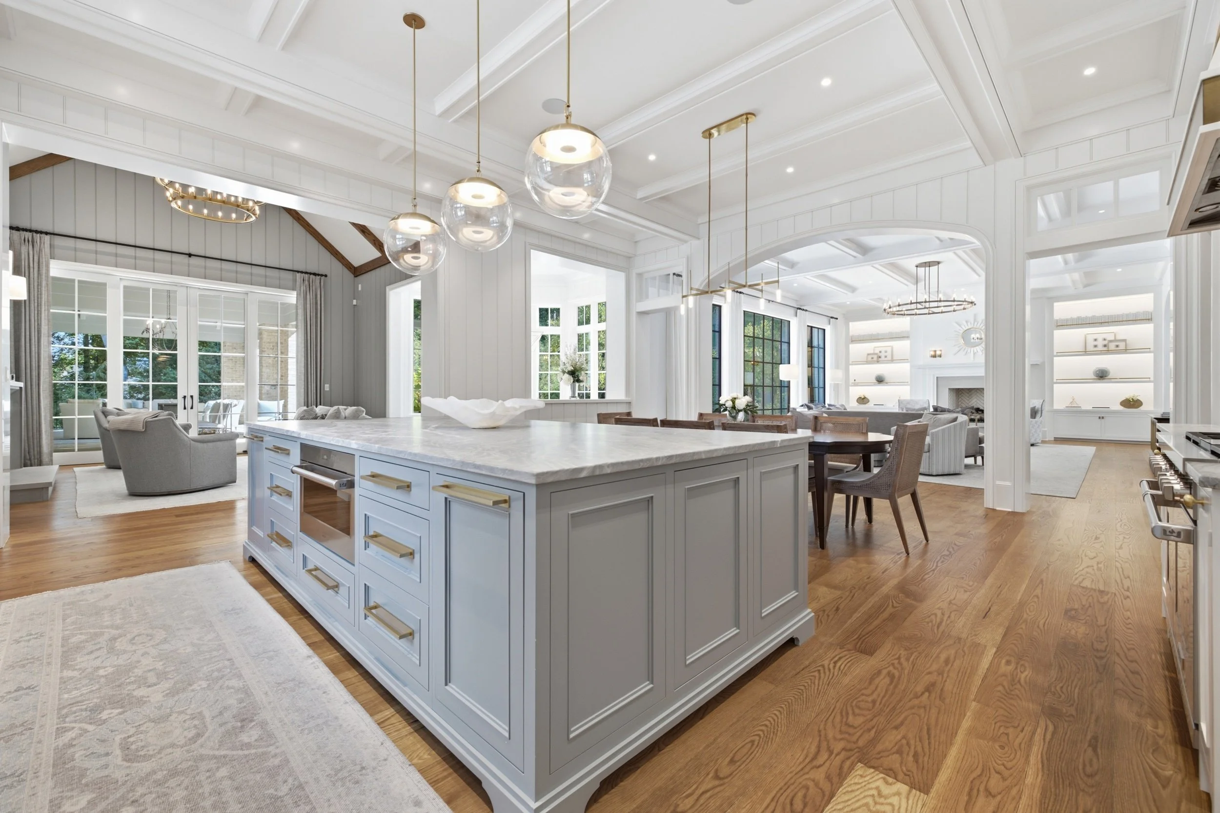 Bright open kitchen and living room with white walls, large windows, wooden floors, modern lighting fixtures, and a kitchen island with a white marble top.