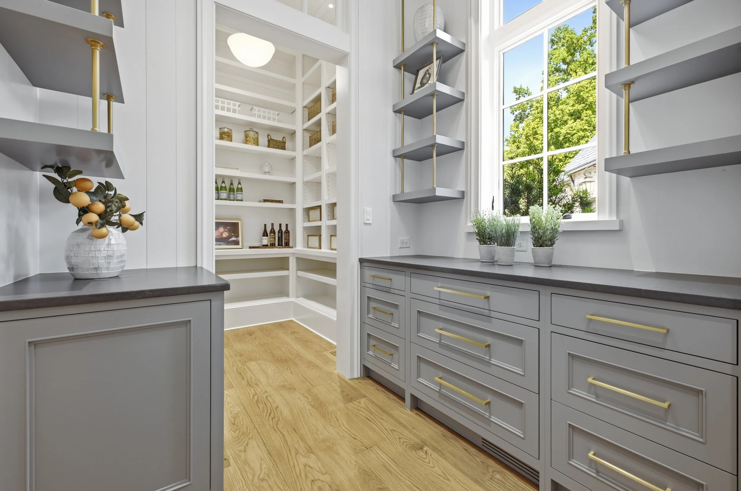 A bright, modern kitchen corner with white cabinetry, light wood flooring, and decorative plants on the counter near a large window showcasing green trees outside.