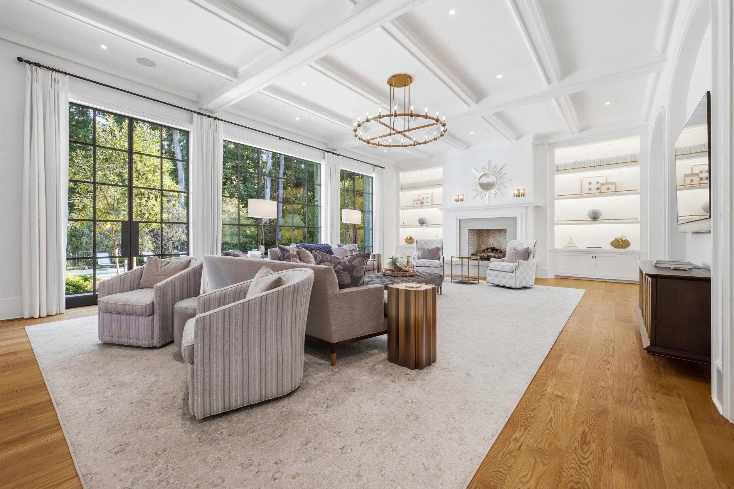 Bright living room with large windows, white walls, and a high tray ceiling. Furnished with a white rug, multiple sofas, and chairs. Elegant gold and wood chandelier and built-in shelves with decorative items surround a fireplace.