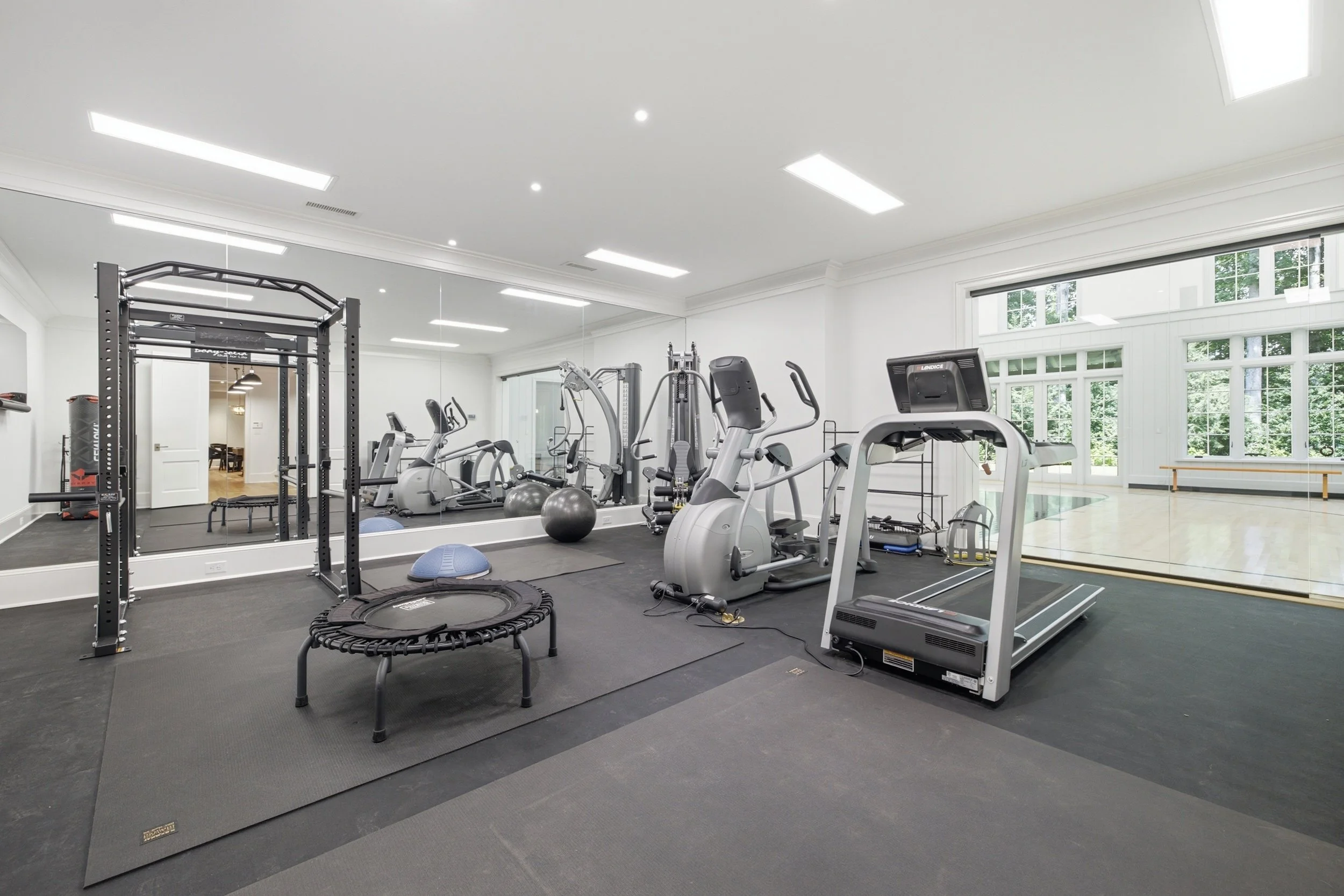 A modern home gym with various exercise equipment including treadmills, stationary bikes, free weights, stability balls, and a mini trampoline, with large windows and a mirror on the wall.