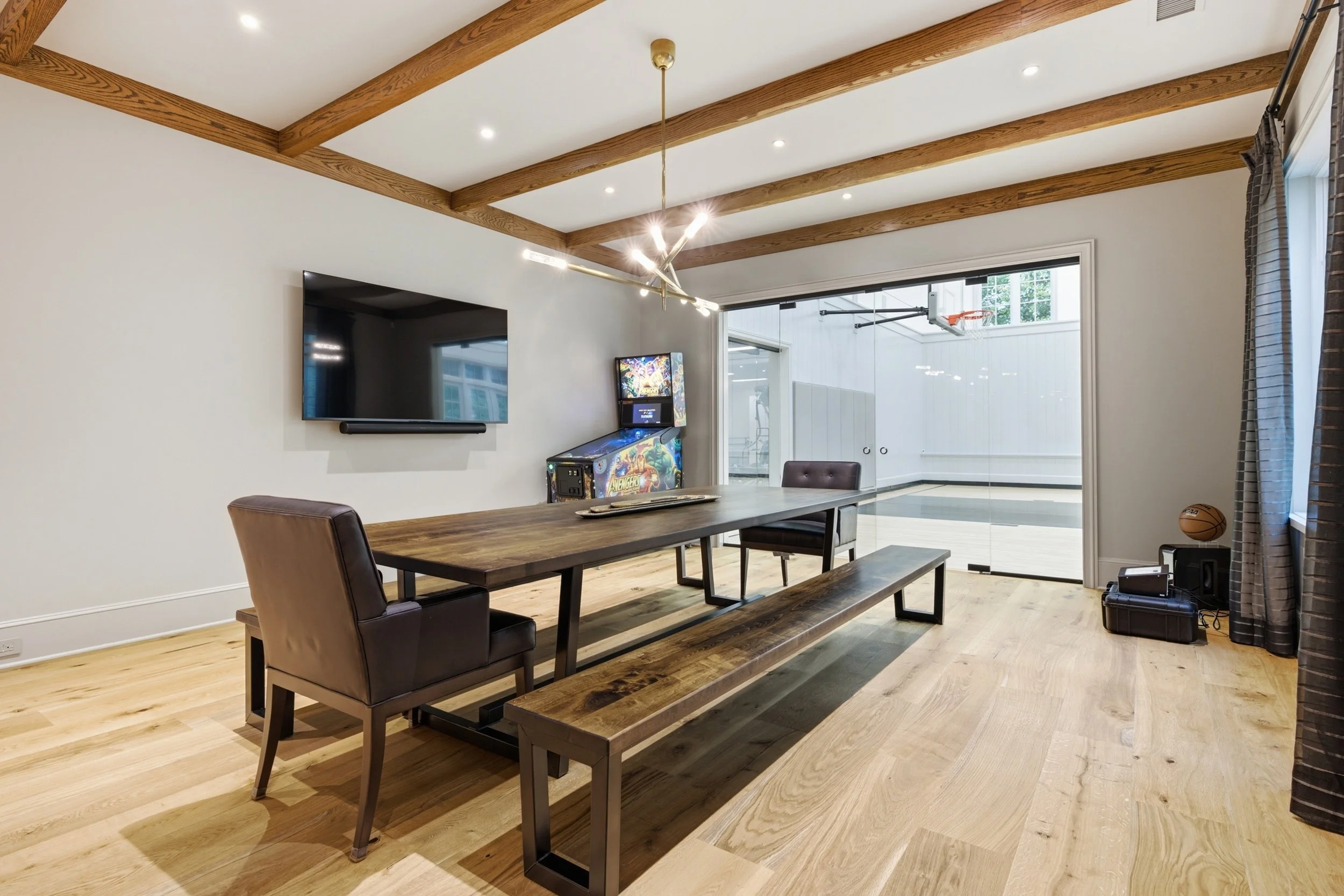 Living room with wooden beams on ceiling, wall-mounted TV, arcade game, dining table with chairs, sliding glass door, basketball, and a basketball hoop area.