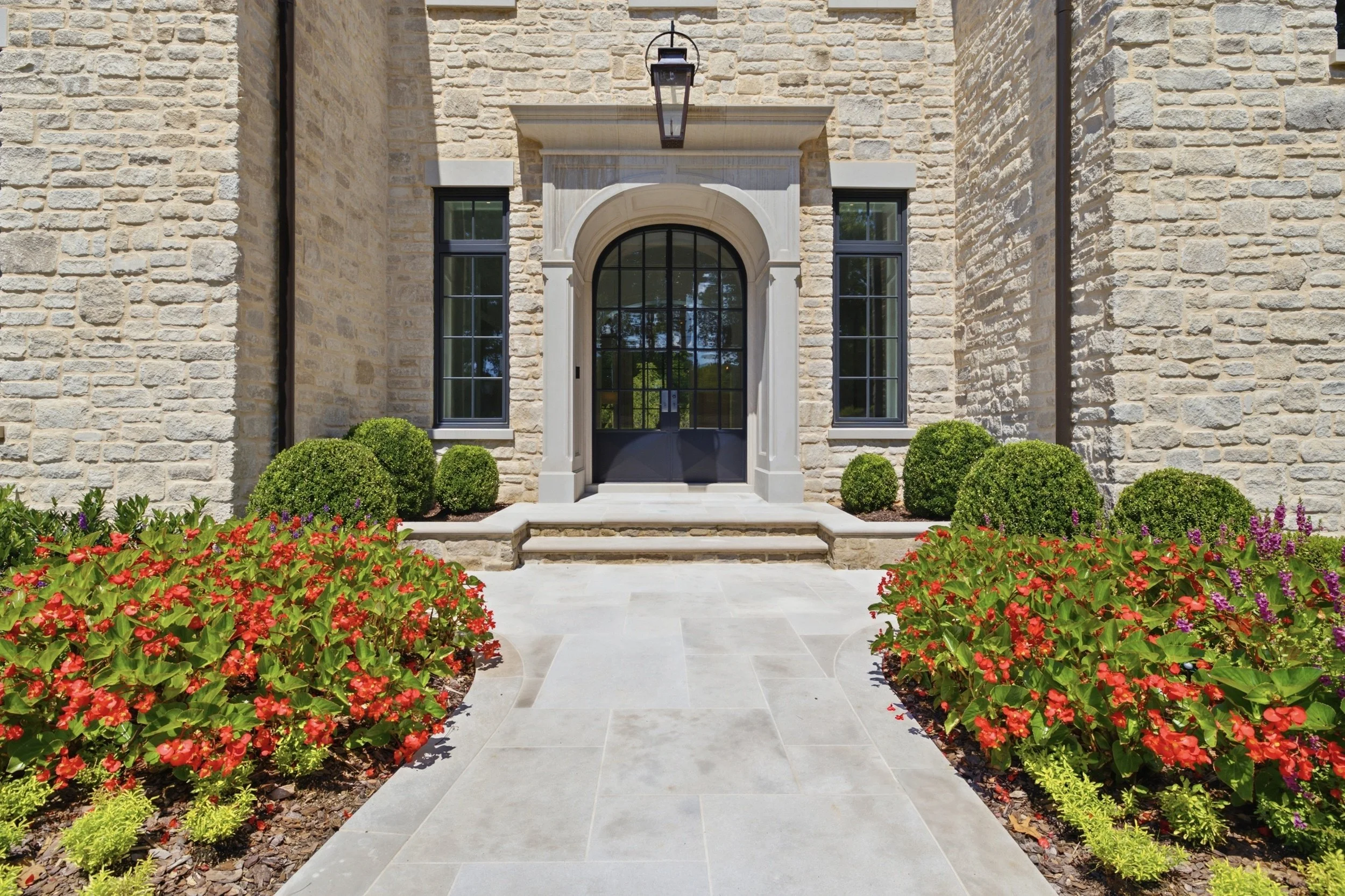 Front entrance of a home with stone exterior walls, black framed doors and windows, a stone staircase, and colorful flower beds with bushes in front.