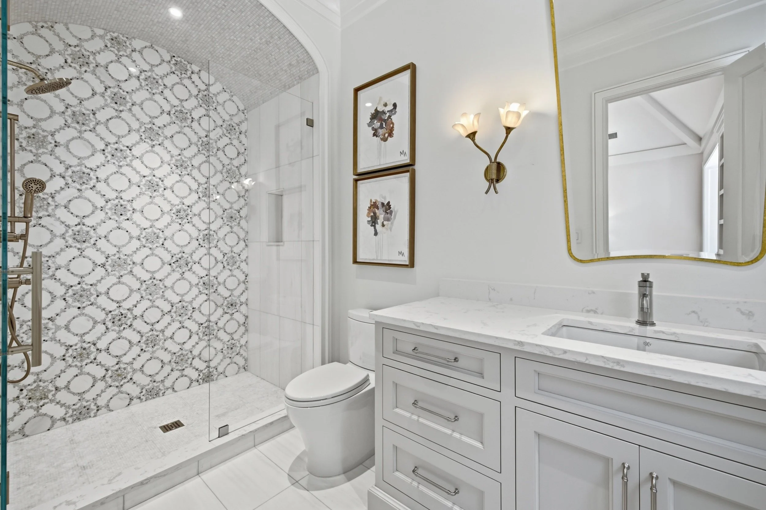 Modern bathroom with shower area separated by glass, decorative tiles, white vanity with marble countertop, framed artwork, and a gold wall-mounted light fixture.