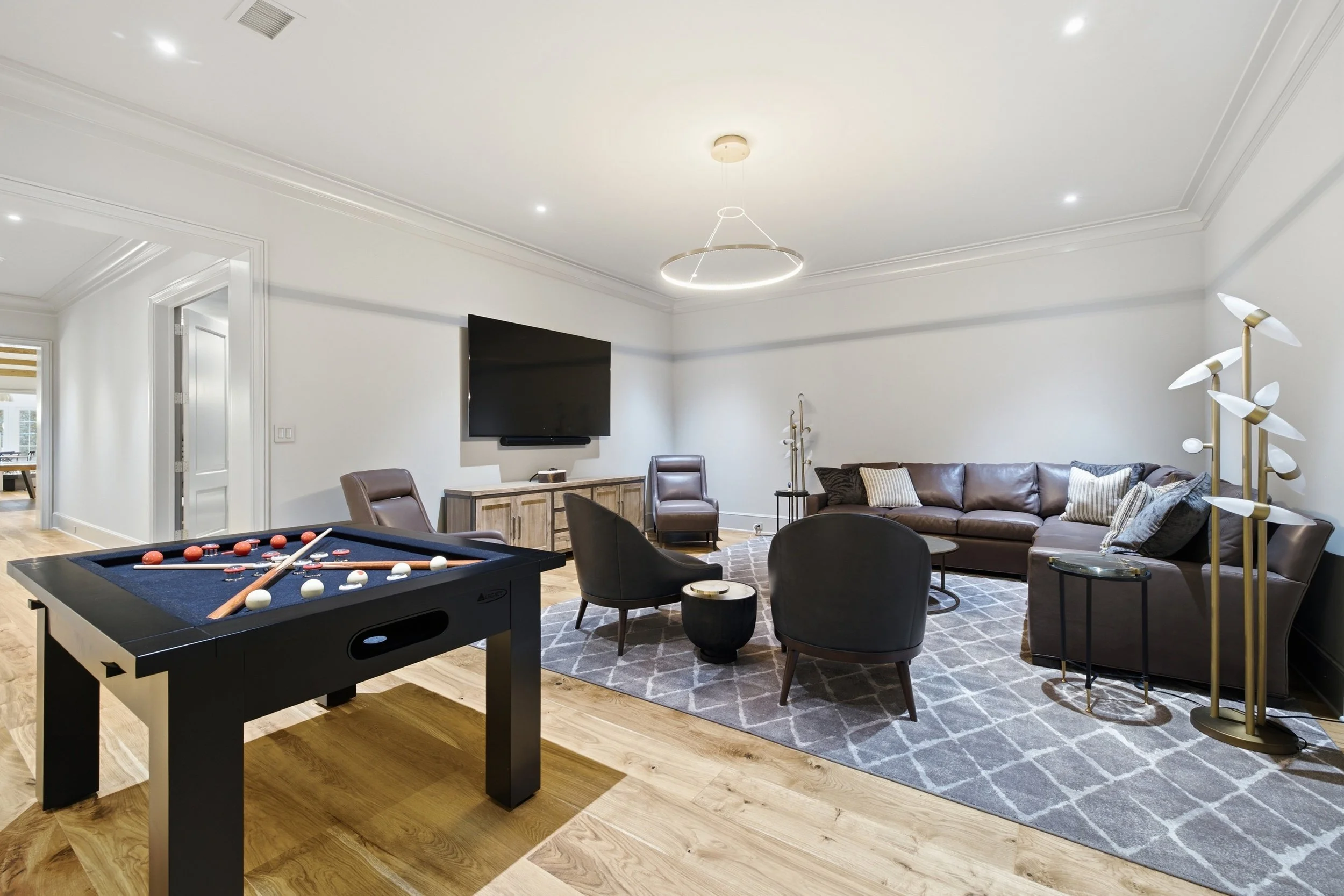 Living room with a foosball table, modern seating including a corner leather sofa with pillows, armchairs, a patterned rug, a wall-mounted TV, modern lighting fixtures, and neutral decor.