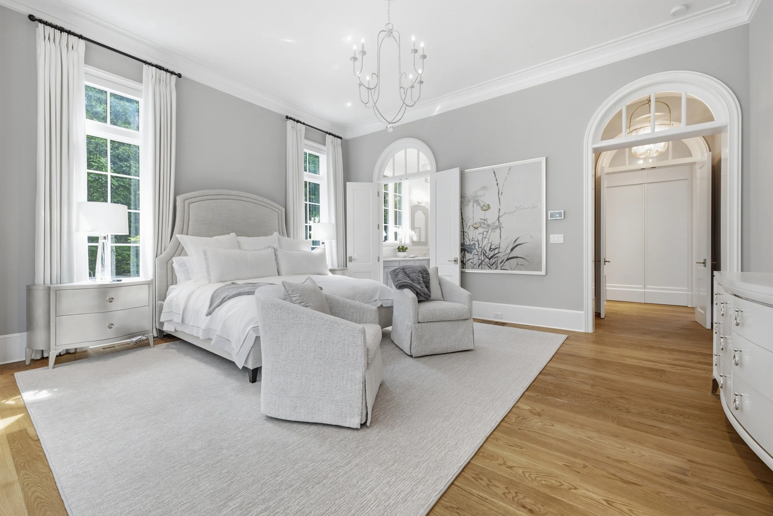 A bright, elegant bedroom with large windows, white drapes, a chandelier, a white upholstered bed, a sitting area with a beige armchair, a large gray rug, and hardwood floors.