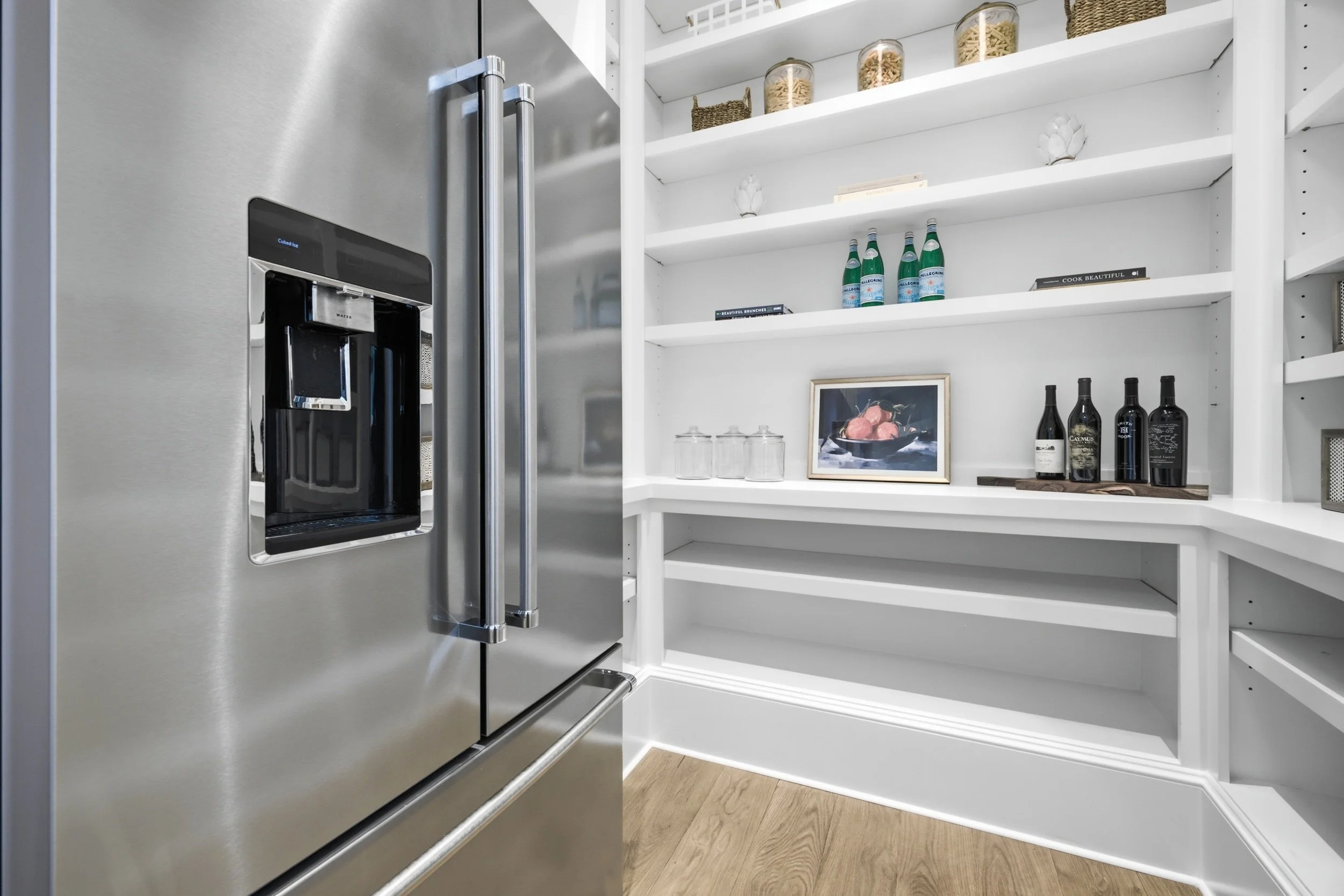 Stainless steel refrigerator next to white open shelving with glass jars, bottles, books, and framed picture inside a kitchen.