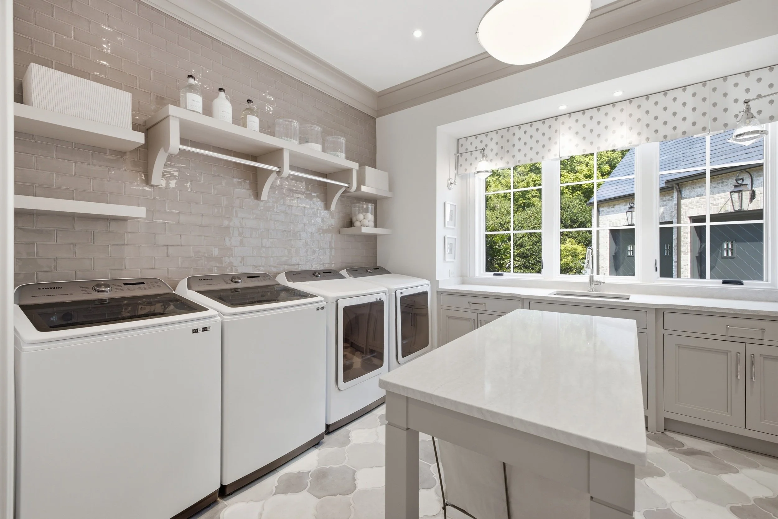 A laundry room with a row of washing machines and a white countertop island near a window with a polka dot valance. There are shelves with bottles and jars on the beige brick wall and decorative framed pictures on the white wall. Large windows reveal