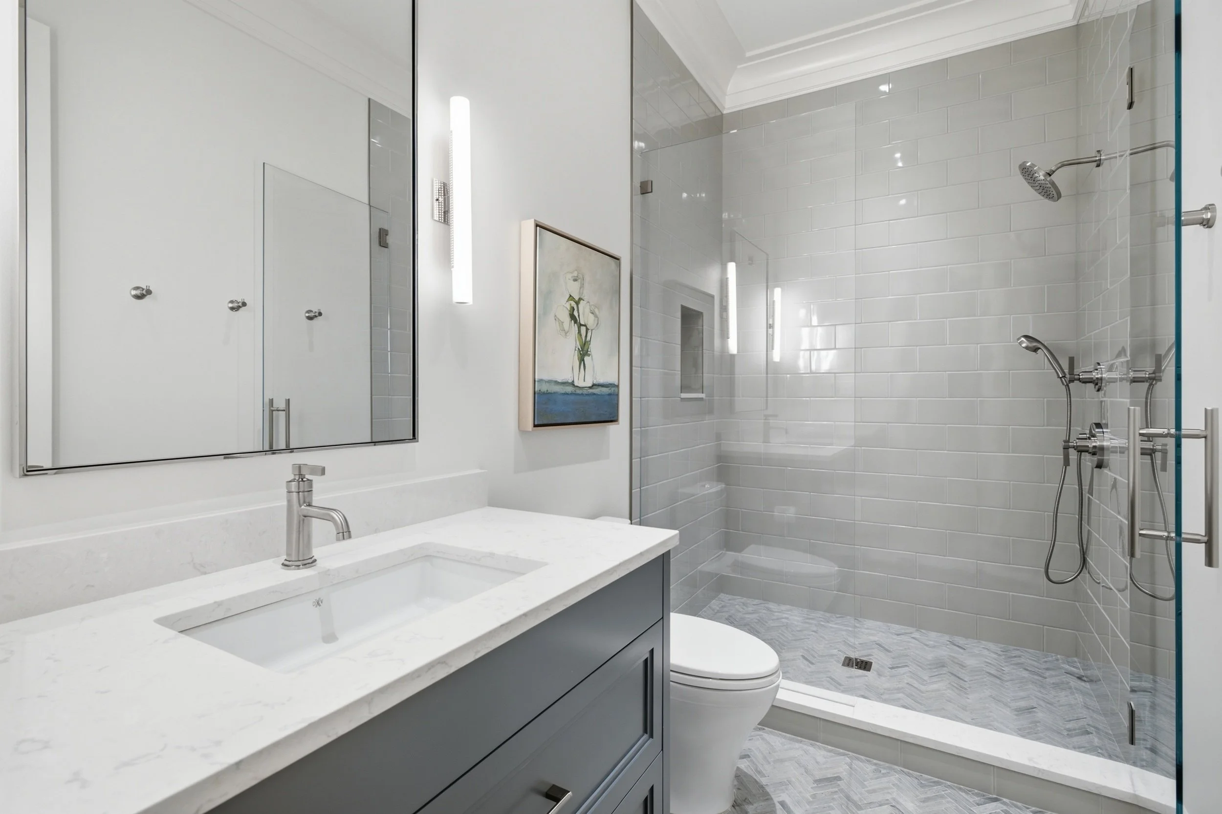 Modern bathroom featuring a white vanity with a marble countertop, a mirror, a blue cabinet below, wall-mounted lights, a framed artwork, a toilet, and a glass-enclosed shower with white subway tiles and multiple showerheads.