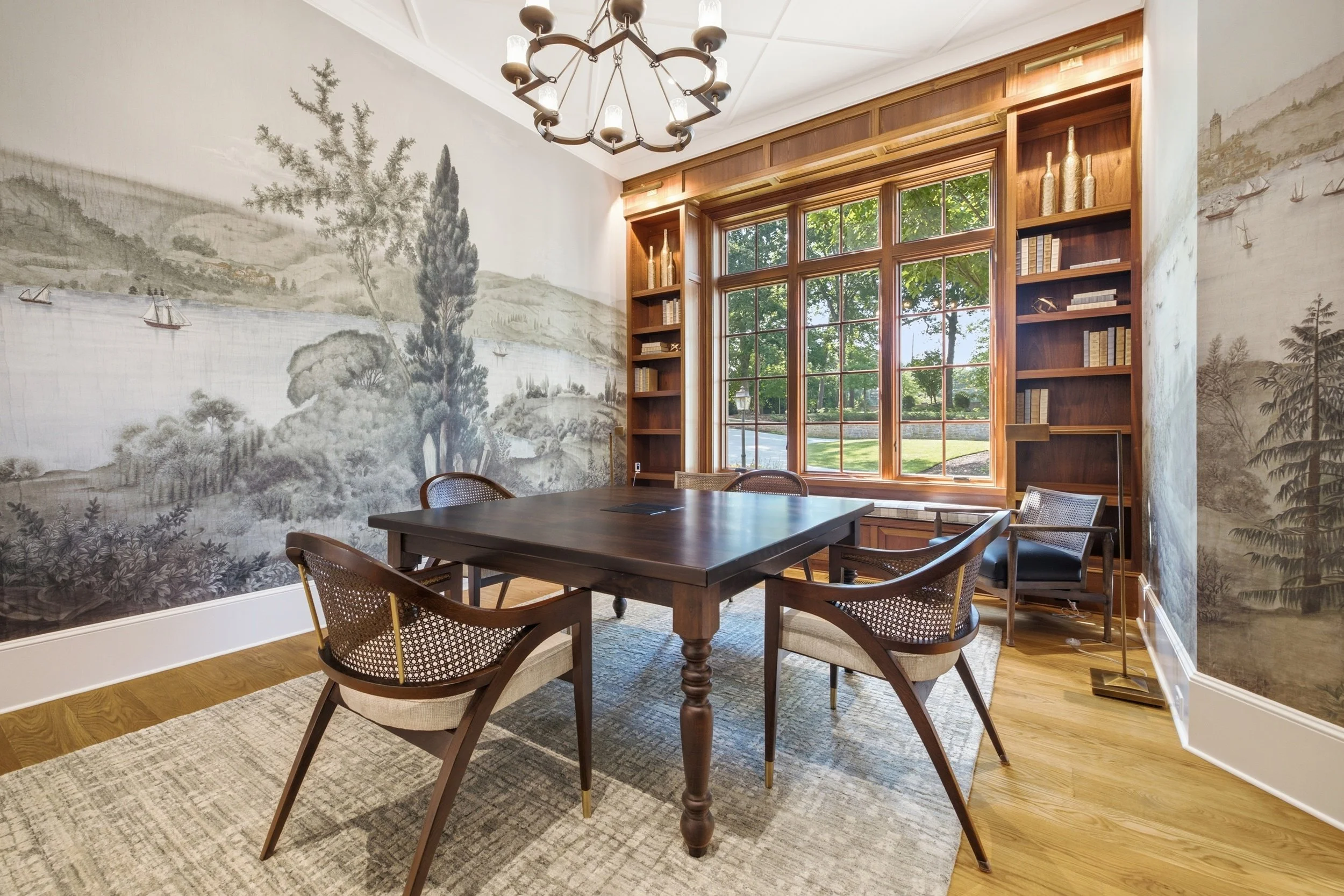 A well-lit dining or meeting room with a large wooden table surrounded by wooden chairs with cane backs. The room features large windows with wooden frames, allowing natural light and offering a view of trees outside. One wall has a mural or wallpape