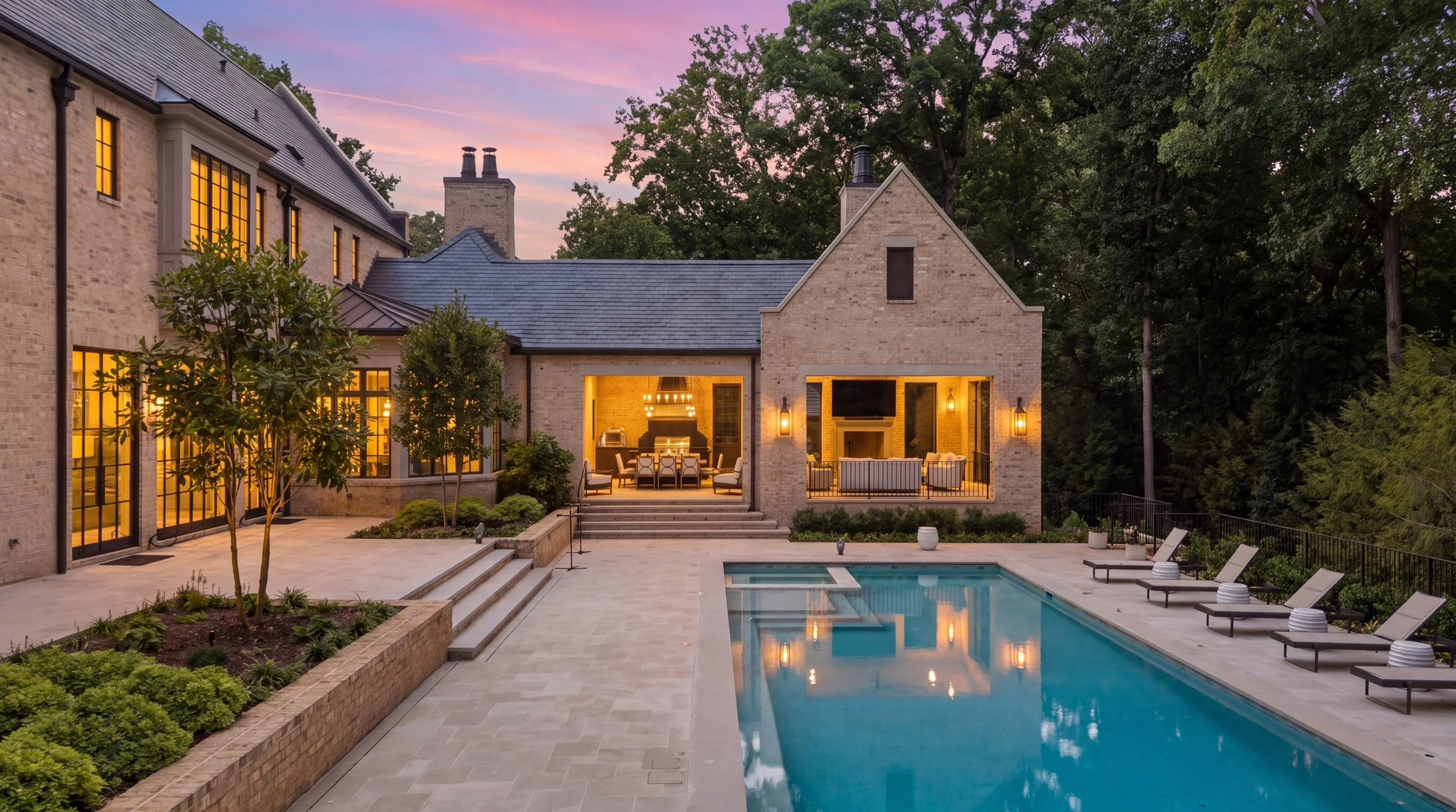 A luxurious backyard with a swimming pool, outdoor seating, and a stone house with large windows illuminated from inside at dusk.