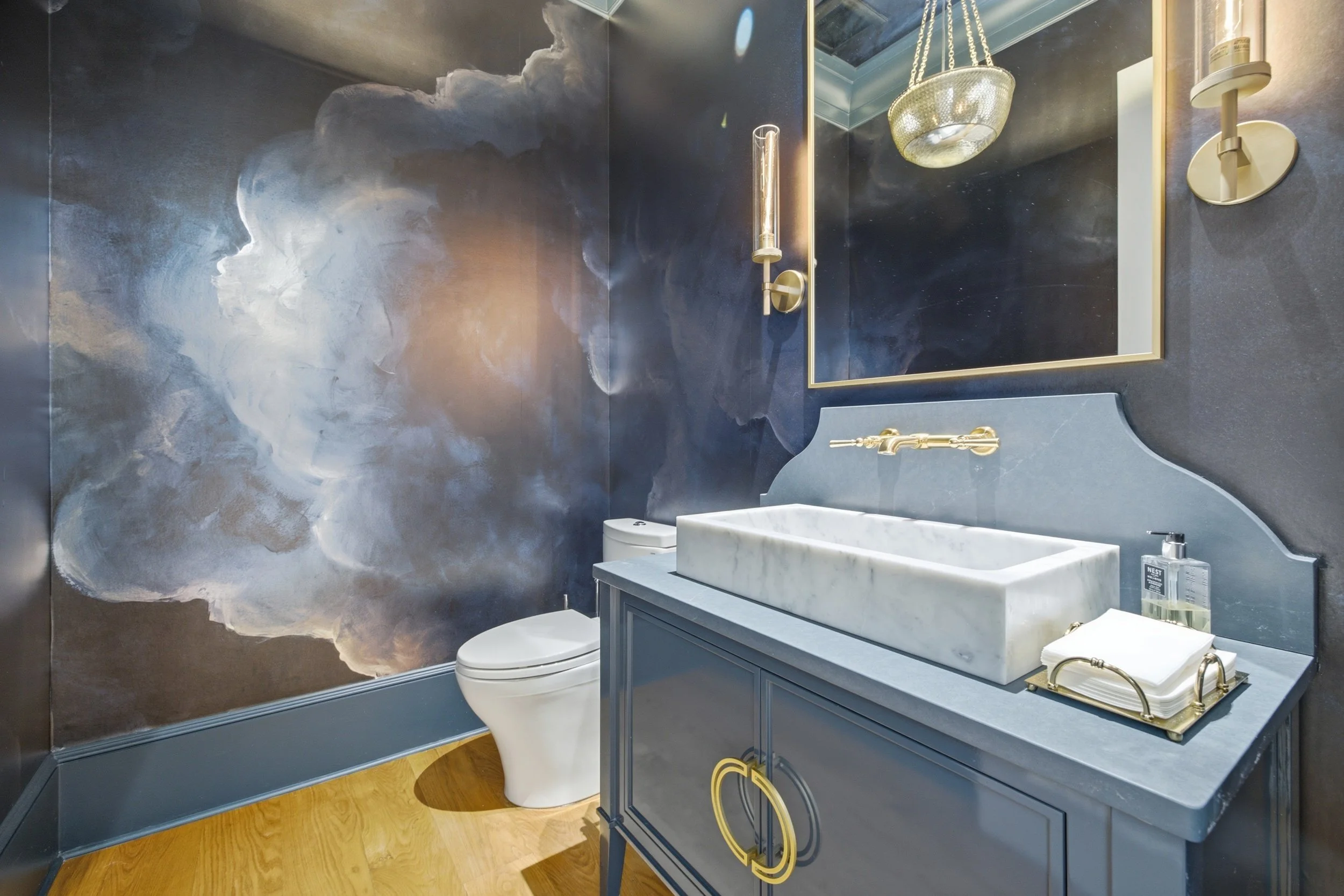 A bathroom with a black and gold theme, featuring a dark cloud-patterned wall, a large mirror with gold trim, a gold chandelier, a marble vanity with gold handles, a modern white toilet, and a wooden floor.