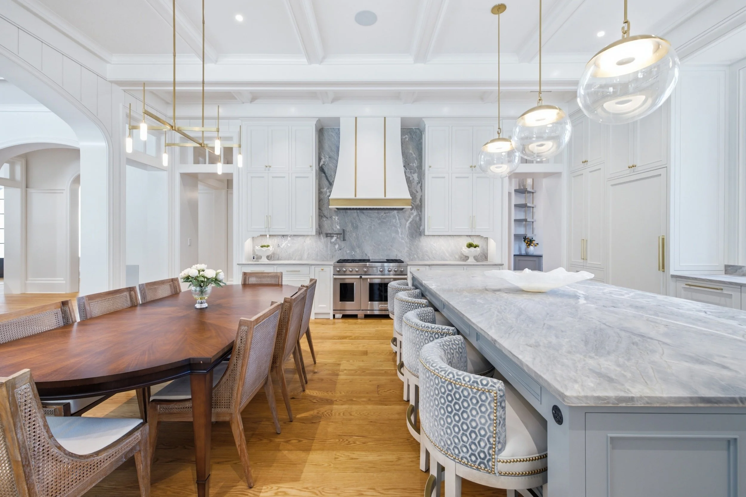 Modern kitchen with white cabinets, marble island, gold lighting fixtures, and wooden dining table with chairs.