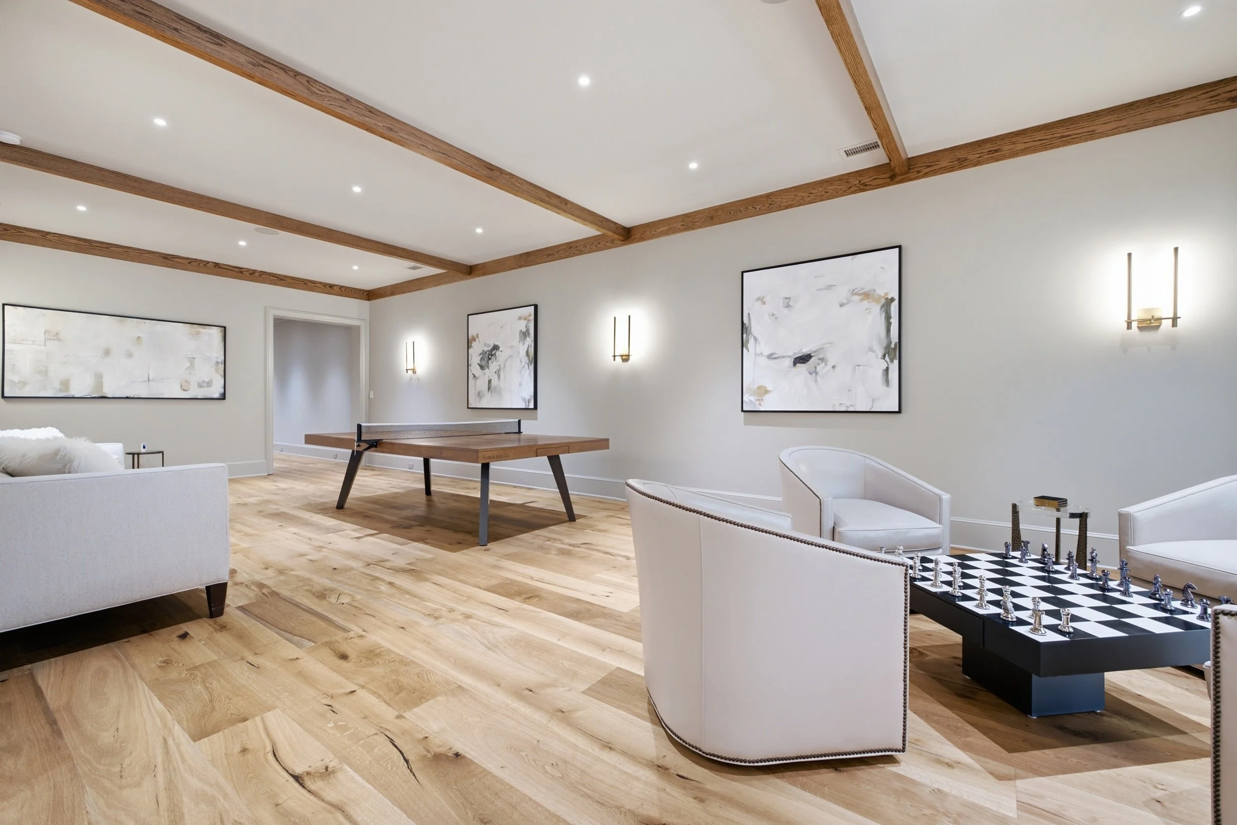 A living room with white walls, wooden beams on the ceiling, and hardwood flooring. It features wall art, a ping pong table, white armchairs, and a chessboard table.