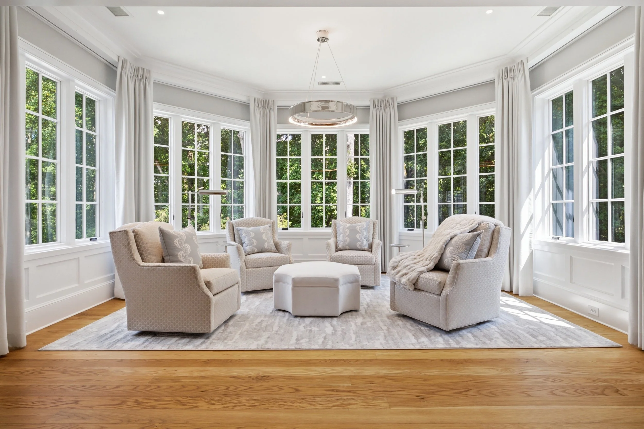 Elegant living room with large bay windows, white curtains, and beige upholstered chairs arranged around a central ottoman, with a view of green trees outside.