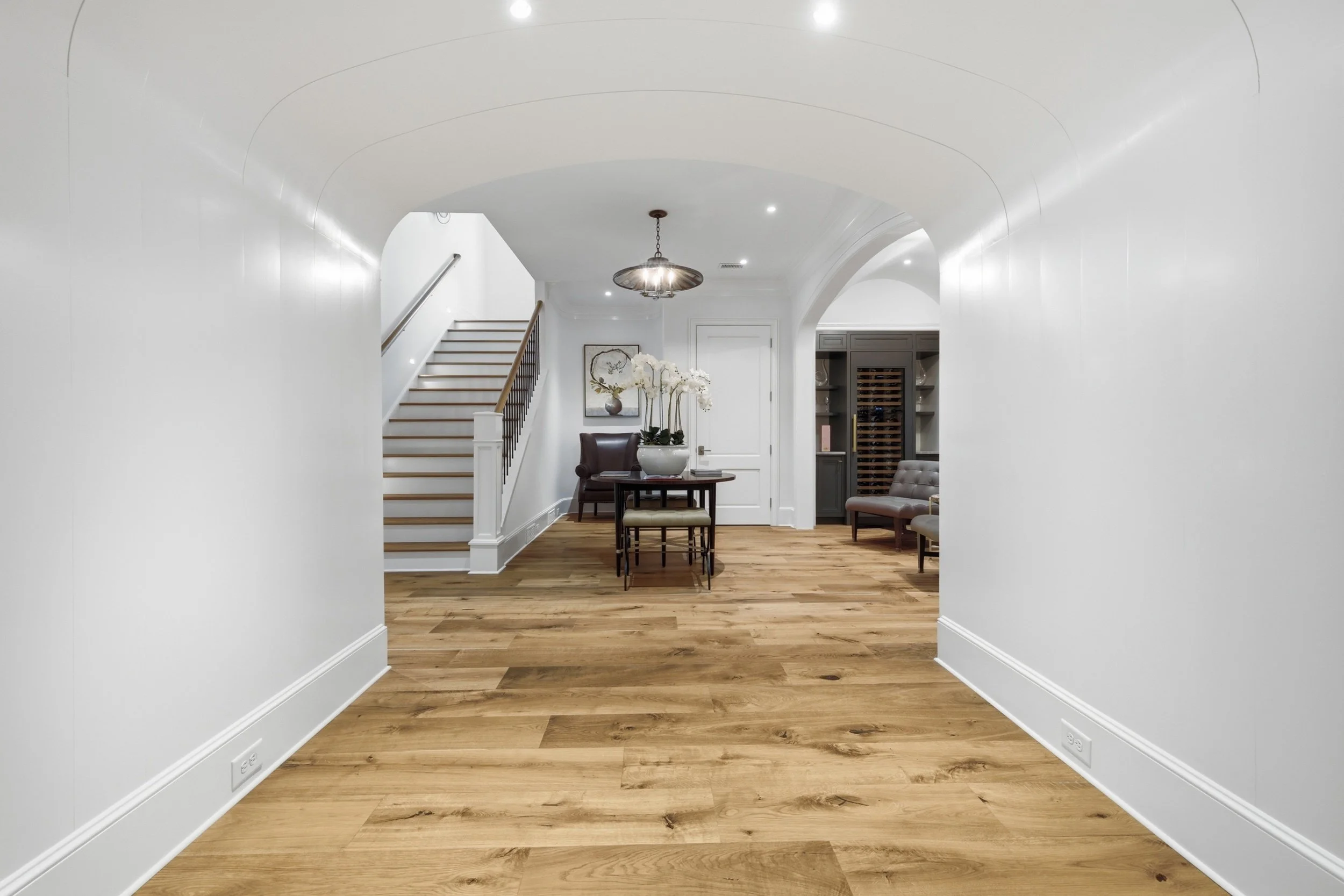 View of a bright, modern hallway with hardwood floors, white walls, and a staircase leading upstairs. The hallway features a small table with a potted orchid and a seating area with chairs, a wall art, and a wine fridge in the background.