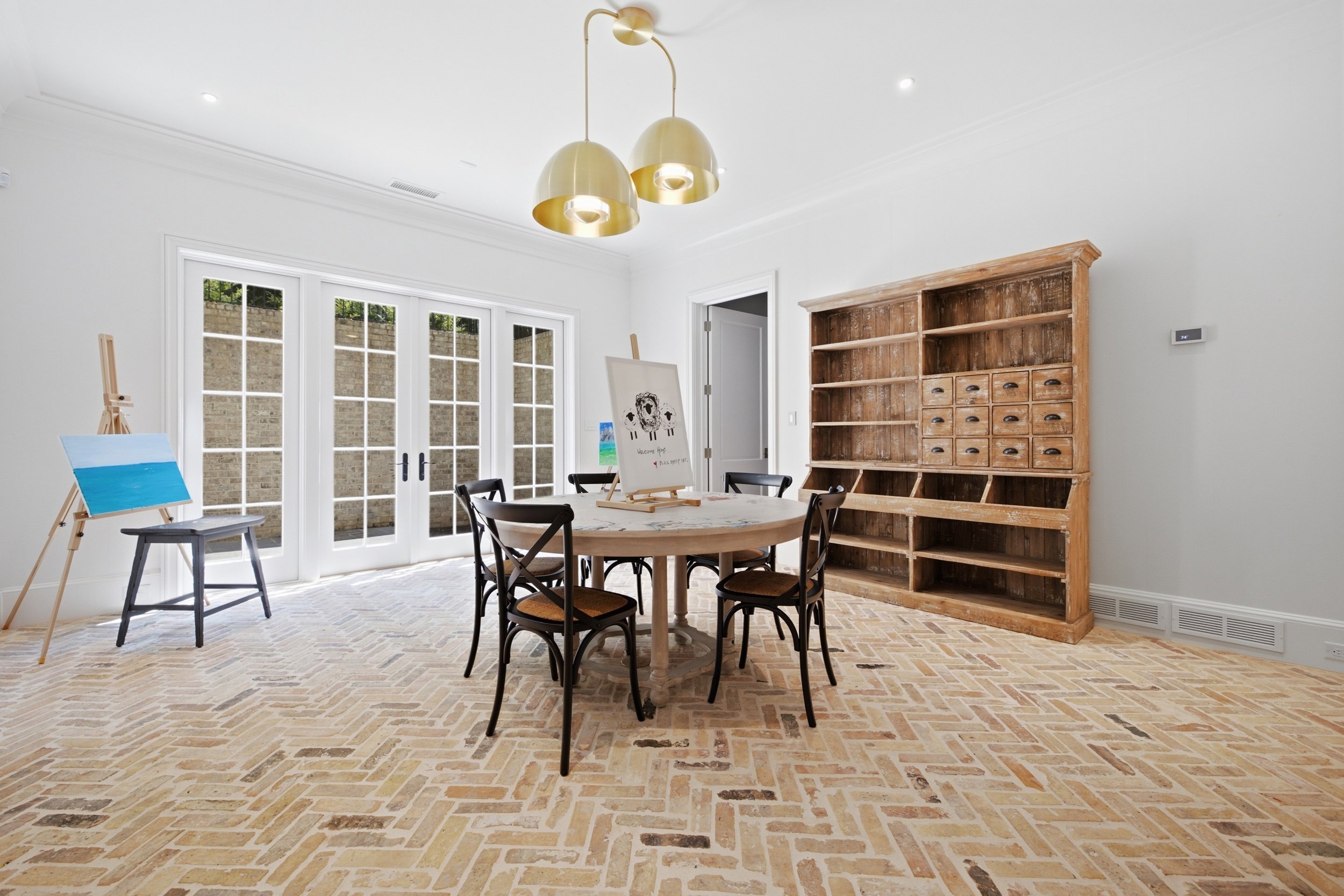 Bright dining room with glass-paneled doors, a round wooden table with four chairs, an easel with painting, a wooden bookshelf, and a modern gold chandelier.
