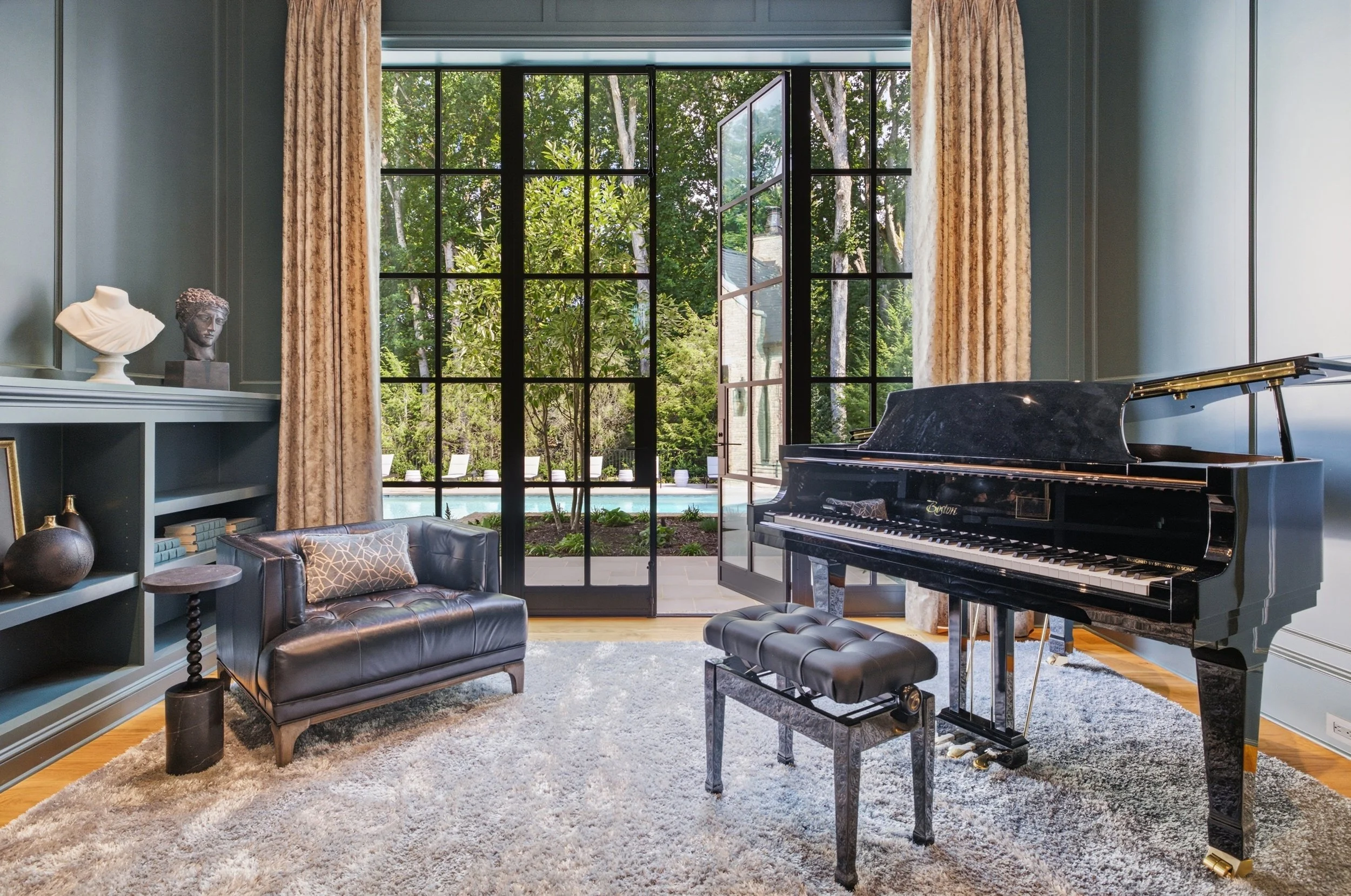 Living room with large windows opening to a patio with pool, featuring a black grand piano, a black leather tufted sofa with a patterned pillow, a gray built-in bookshelf with sculptures and books, and beige curtains.