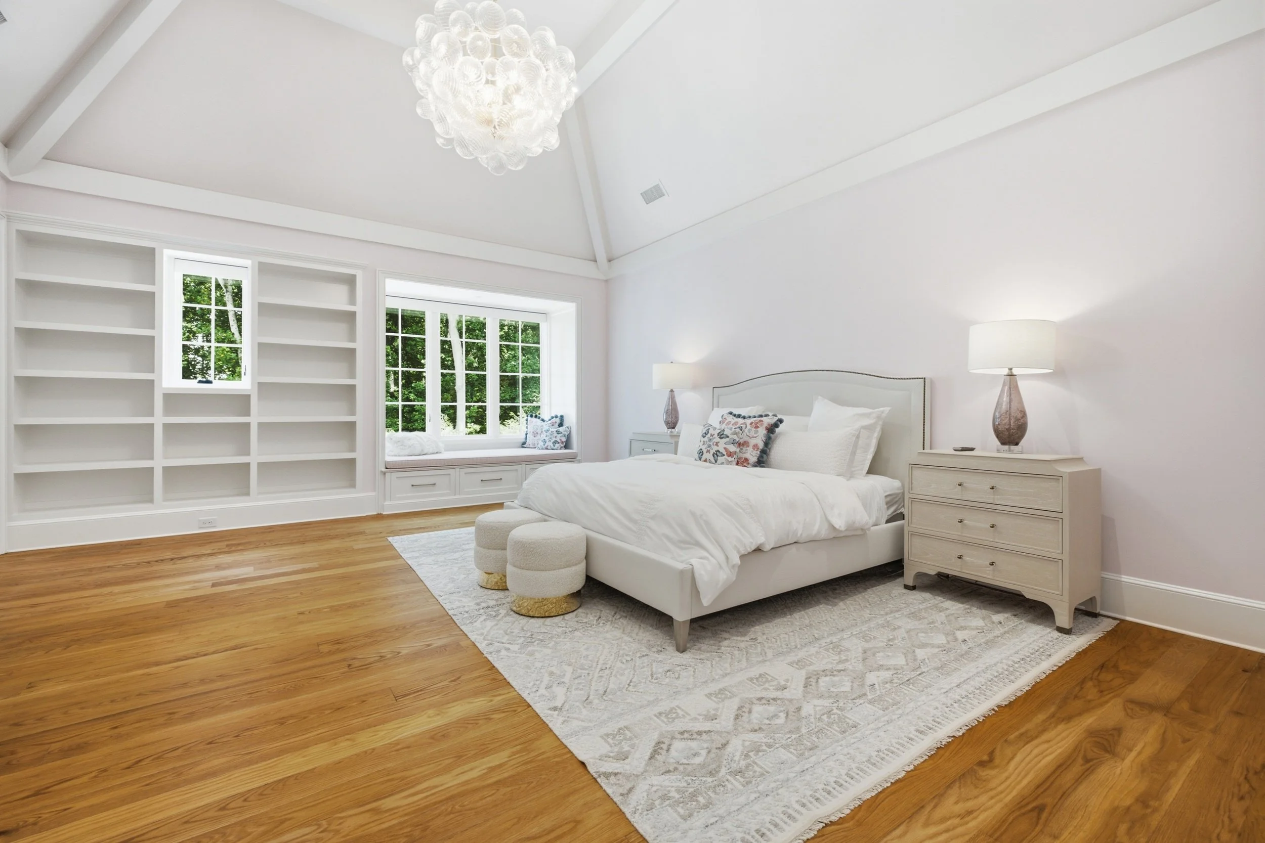 A bedroom with a white bed, two nightstands with lamps, a window seat with pillows, large window panes showing trees outside, a white bookshelf, a ceiling light fixture, white walls, and hardwood flooring.