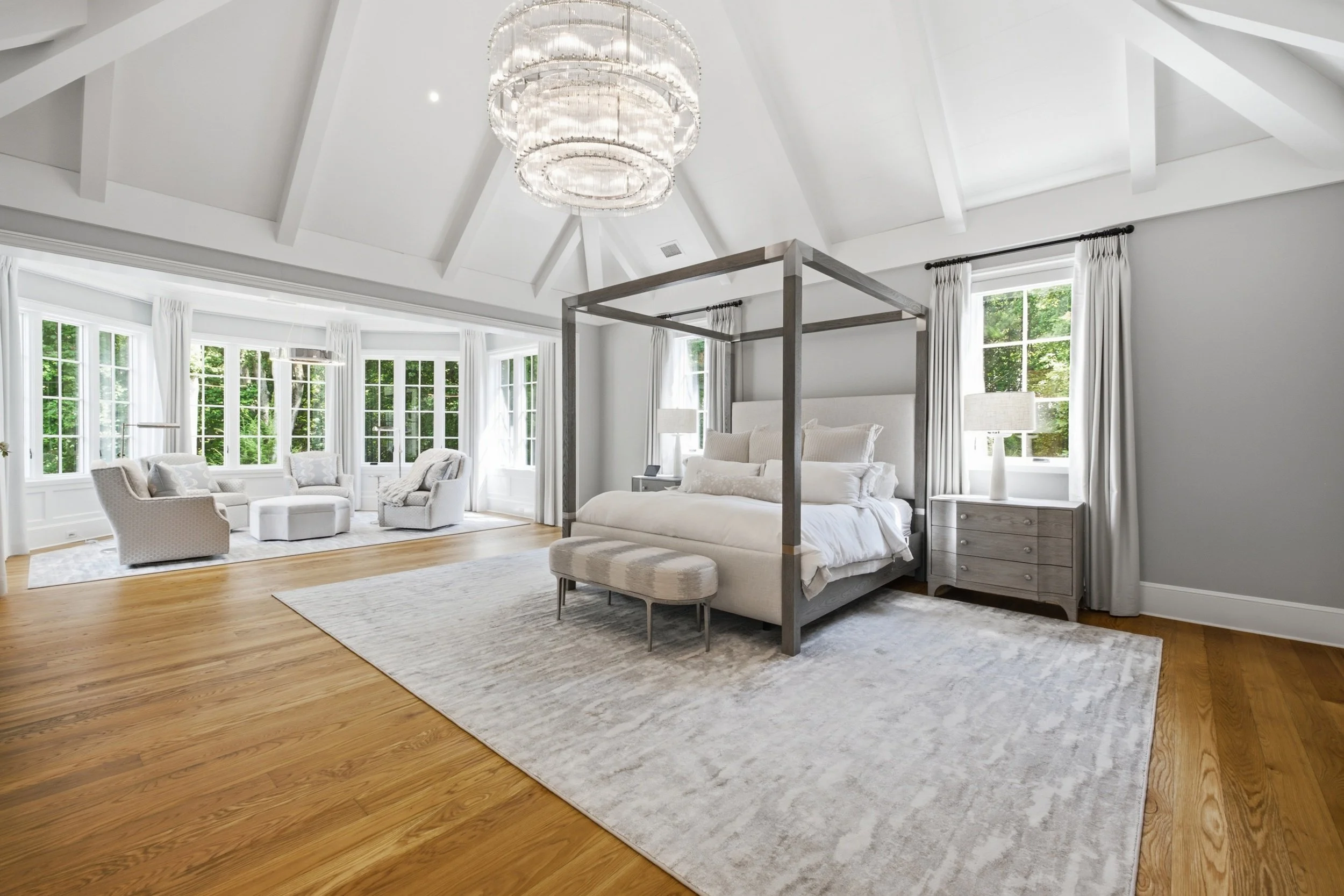 A spacious, bright bedroom with large windows, white walls, and a wooden floor. Features a bed with a canopy frame, a side table, a white floor lamp, and cozy seating area with two armchairs and a round ottoman. A modern chandelier hangs from the whi