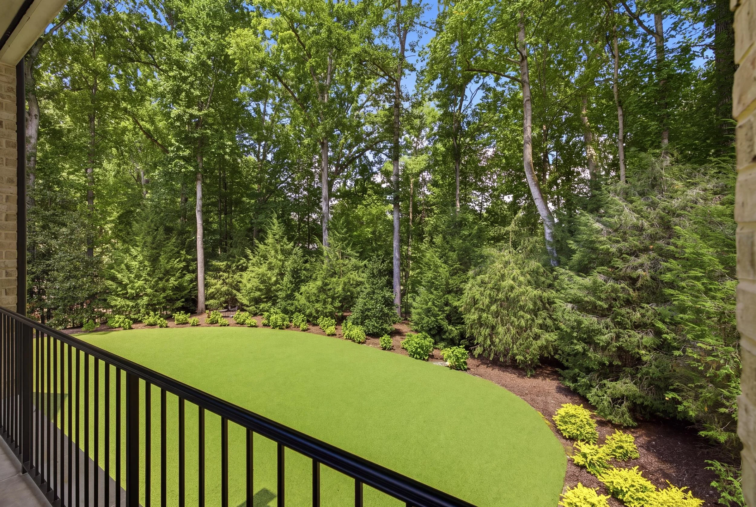 View from a balcony showing a well-maintained lawn, surrounded by lush green trees and bushes, with a dense forest in the background under a bright blue sky.