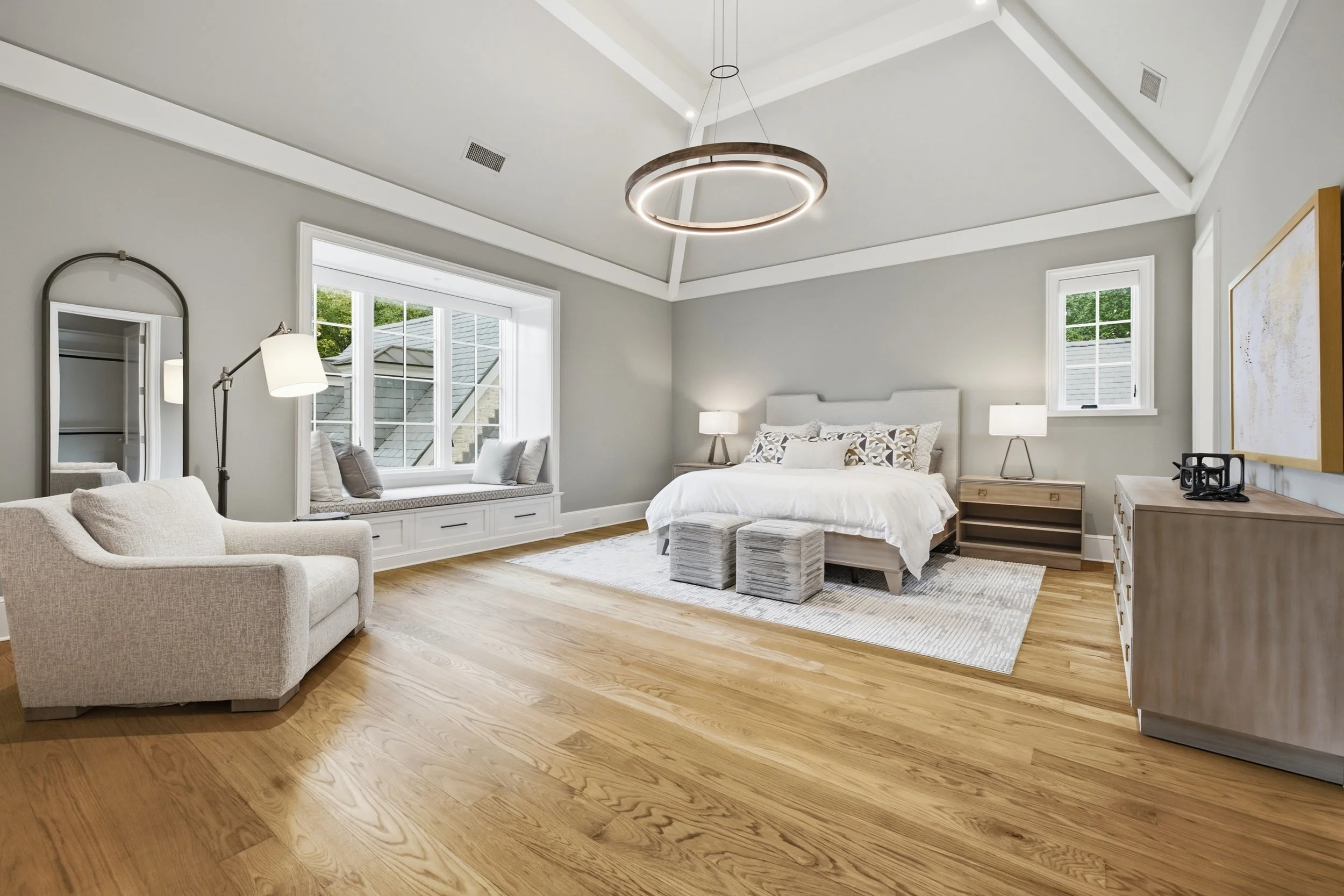 A modern bedroom with a white bed, wooden nightstands with lamps, a beige armchair, and large windows with a view of neighboring rooftops and trees. The ceiling features a circular light fixture and has a vaulted design with white beams.