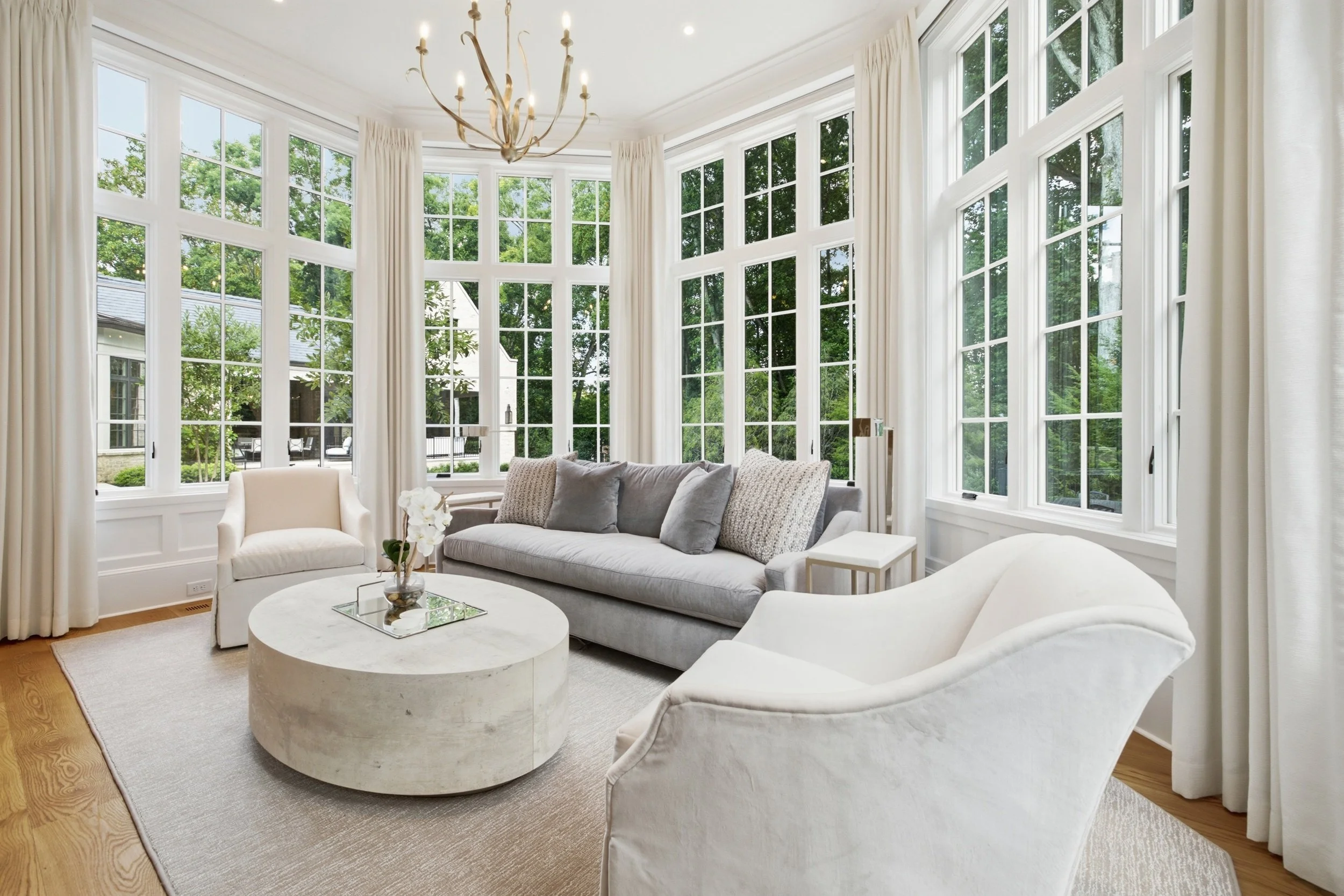Living room with large bay windows, white curtains, a chandelier, cream and gray sofas, a round coffee table with a flower vase, and greenery visible outside.