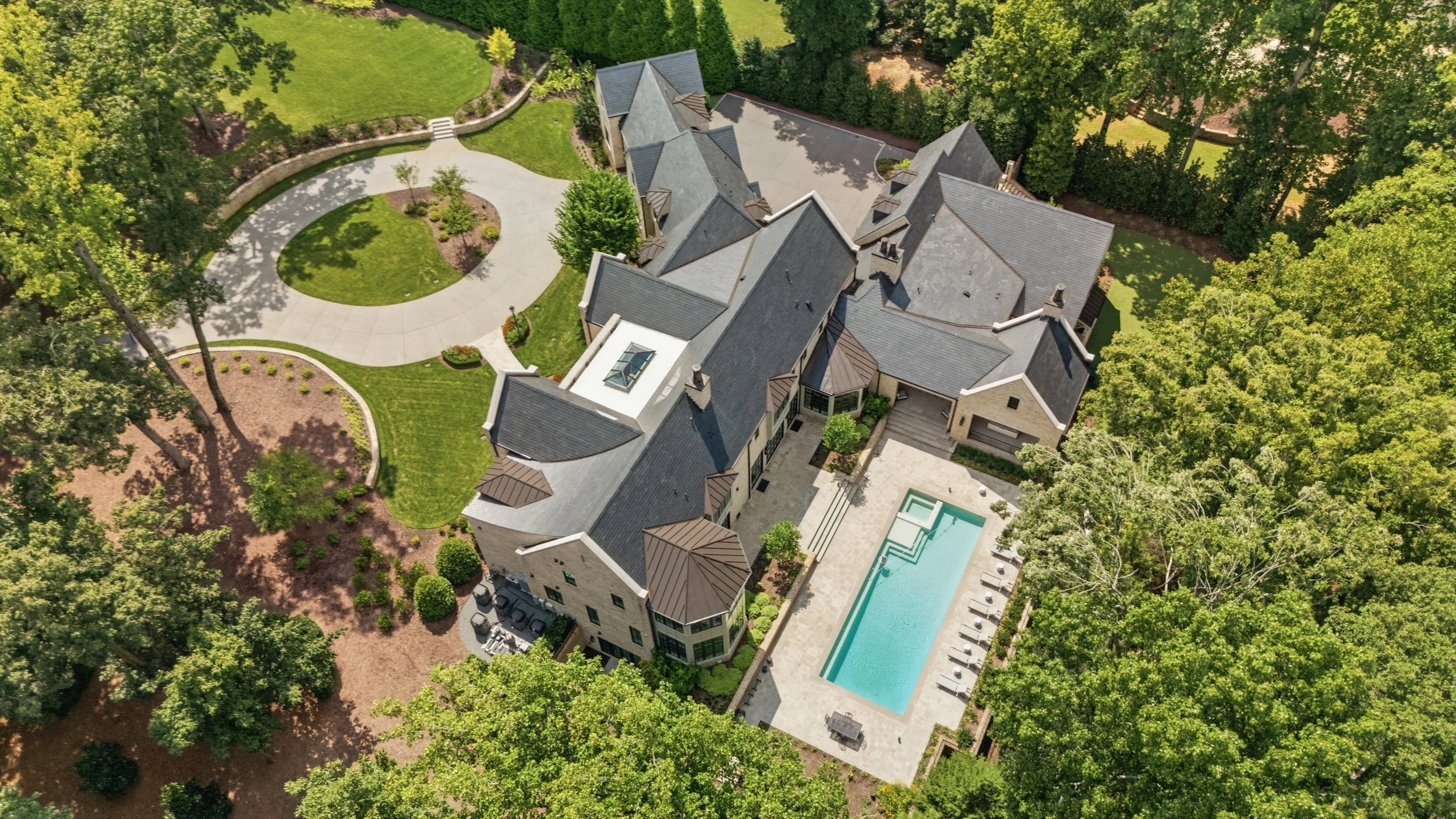 An aerial view of a large, modern house surrounded by green trees and landscaping, including a curved driveway, lawn, and a swimming pool.