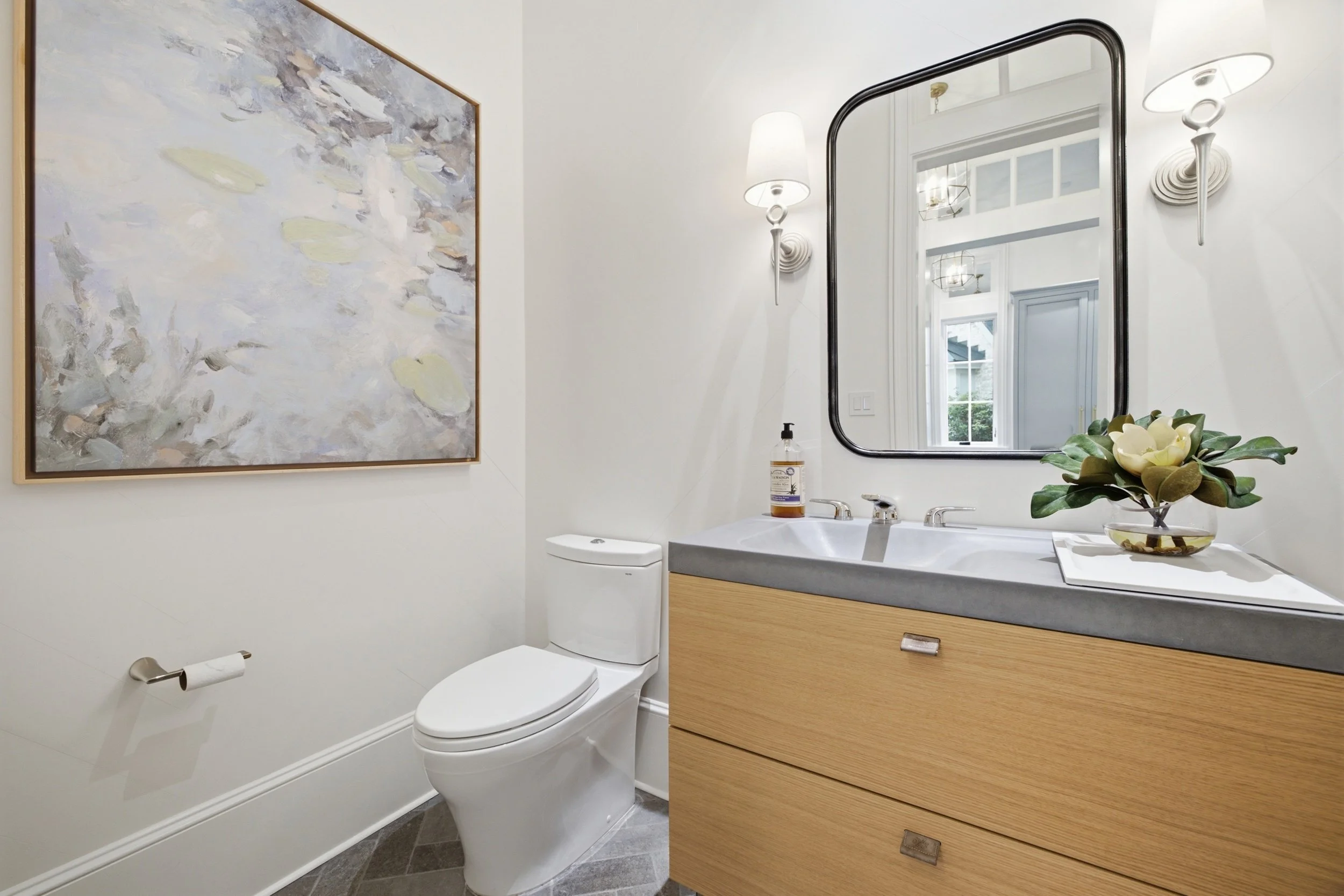 Modern bathroom with white walls, a rectangular mirror, a white toilet, a beige vanity with a gray countertop, a decorative plant, a bottle of hand soap, and wall-mounted light fixtures.