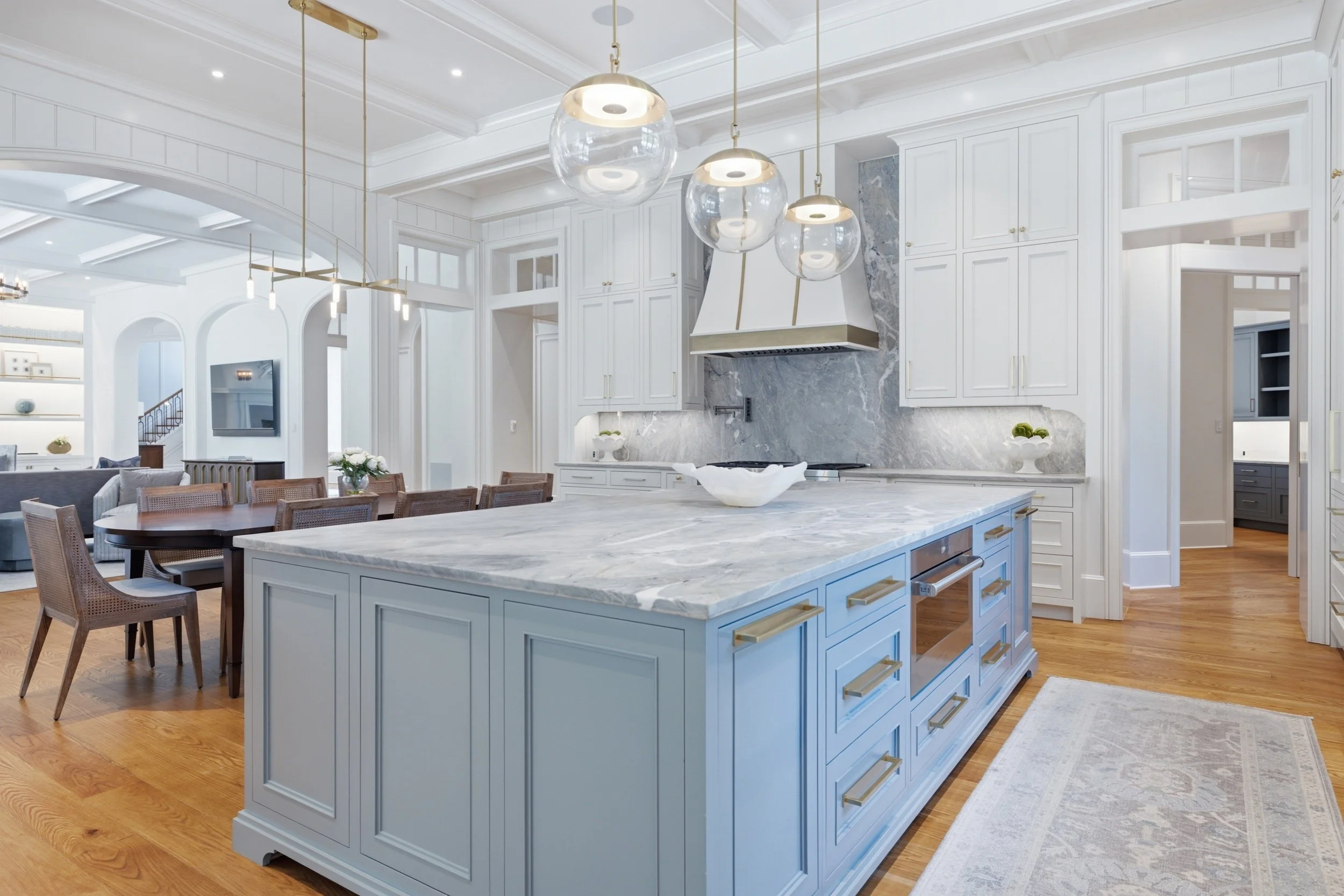 Bright white kitchen with marble island, pendant lights, wooden dining table, and open floor plan connected to living space.