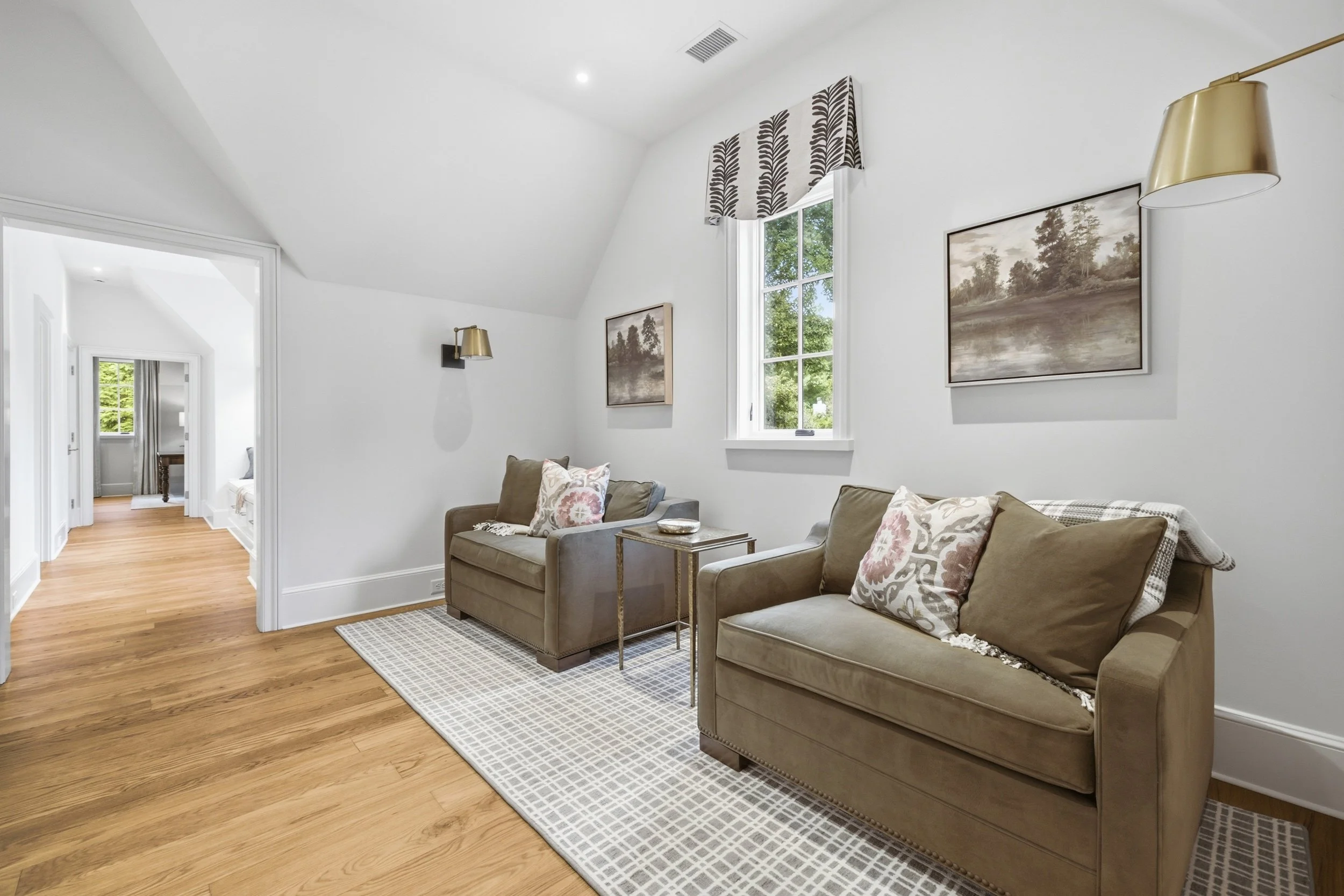 Living room with two beige sofas with floral pillows, a small gold side table, framed landscape paintings, a window with a patterned valance, and wood flooring.