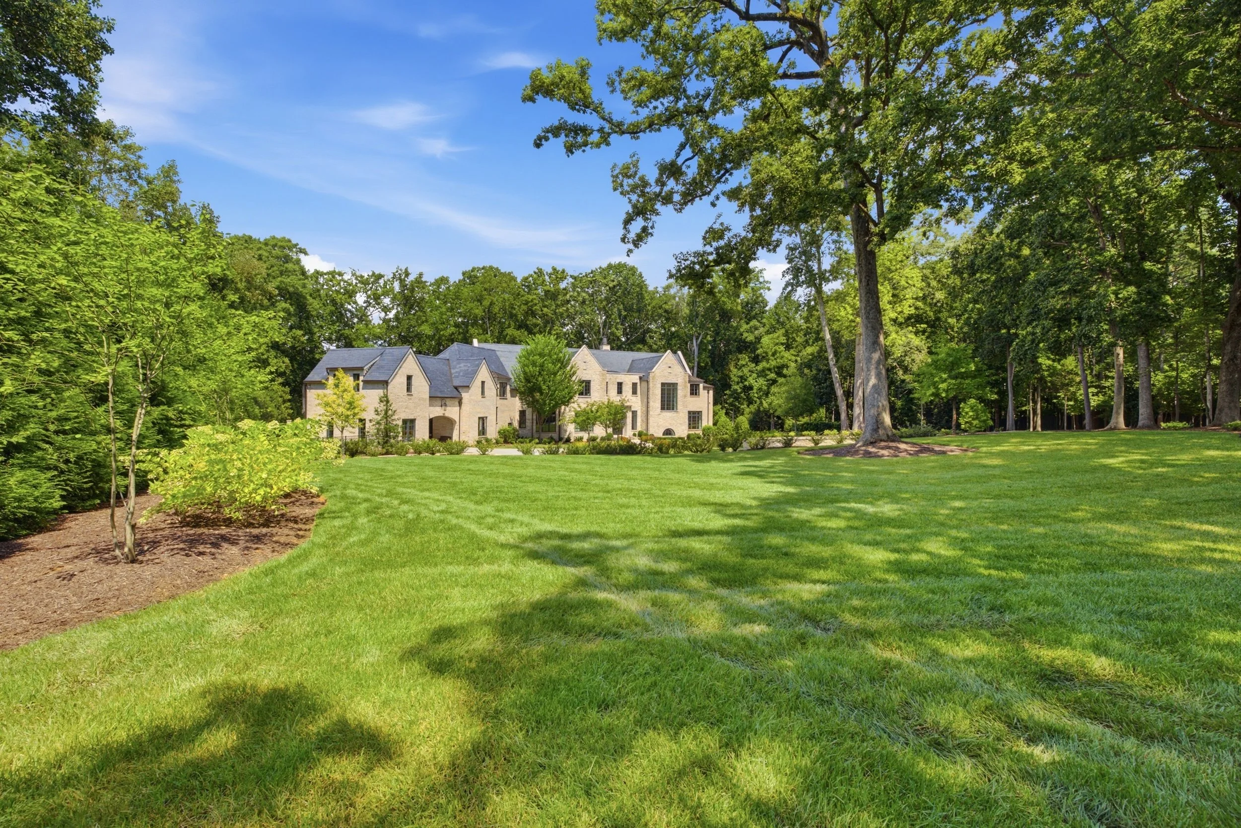 A large house with stone exterior walls, multiple gable roofs, and tall windows, situated in a lush, green, wooded landscape on a bright sunny day with a clear blue sky.