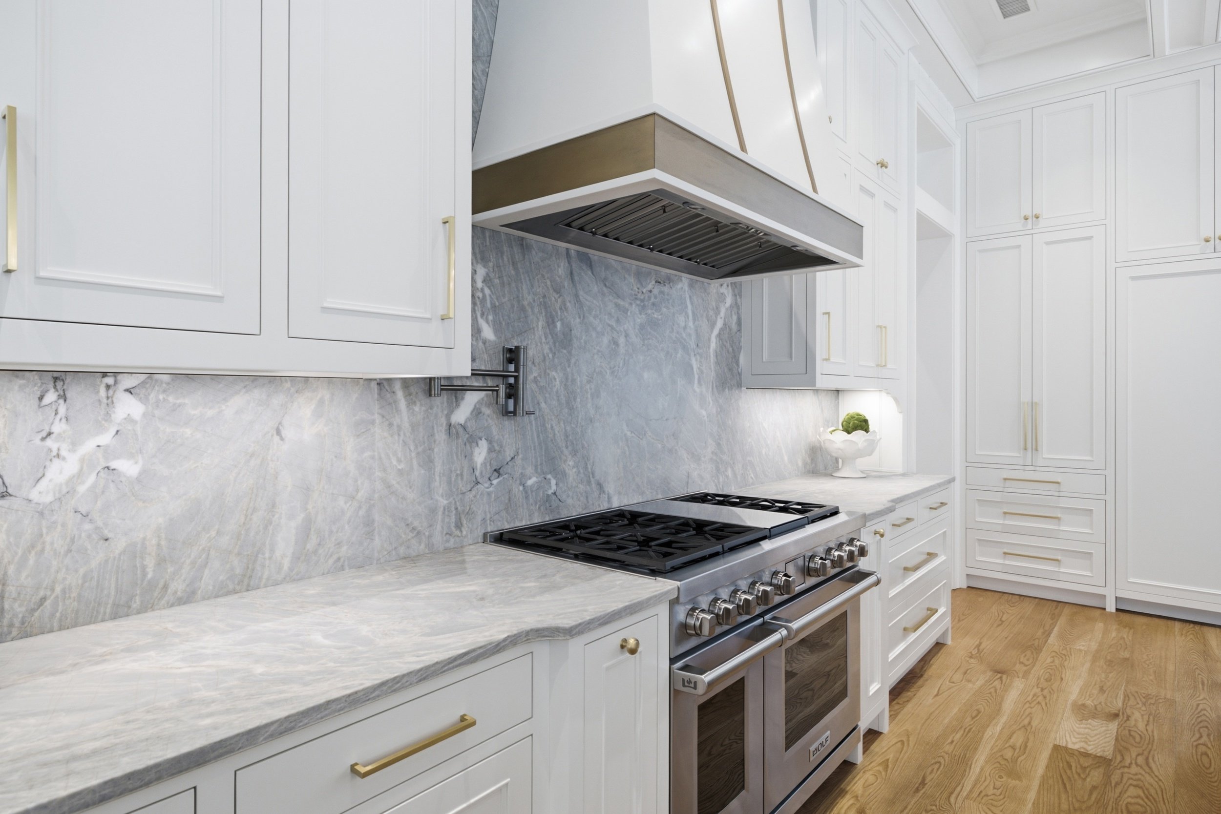 Modern kitchen with white cabinets, marble countertops, and a stainless steel gas stove. The floor is wooden, and there is a potted plant on the counter.
