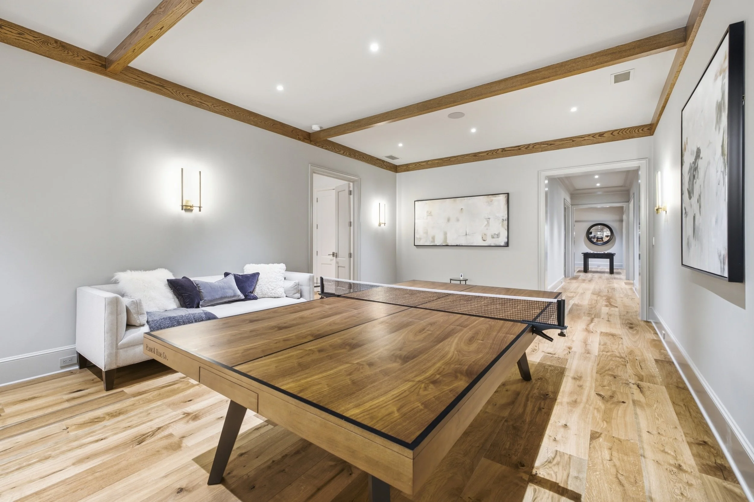 A modern interior living room with a ping pong table, white sofa with cushions, hardwood floors, wooden ceiling beams, and abstract artwork on the walls, with a hallway visible in the background.
