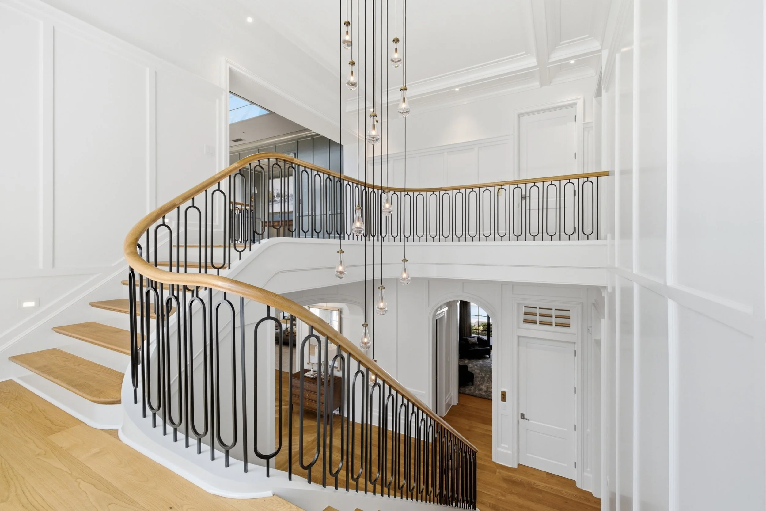 Interior view of a modern staircase with wooden steps, black metal railing, and a curved wooden handrail, in a bright open house with white walls and ceiling, and pendant lights hanging from above.