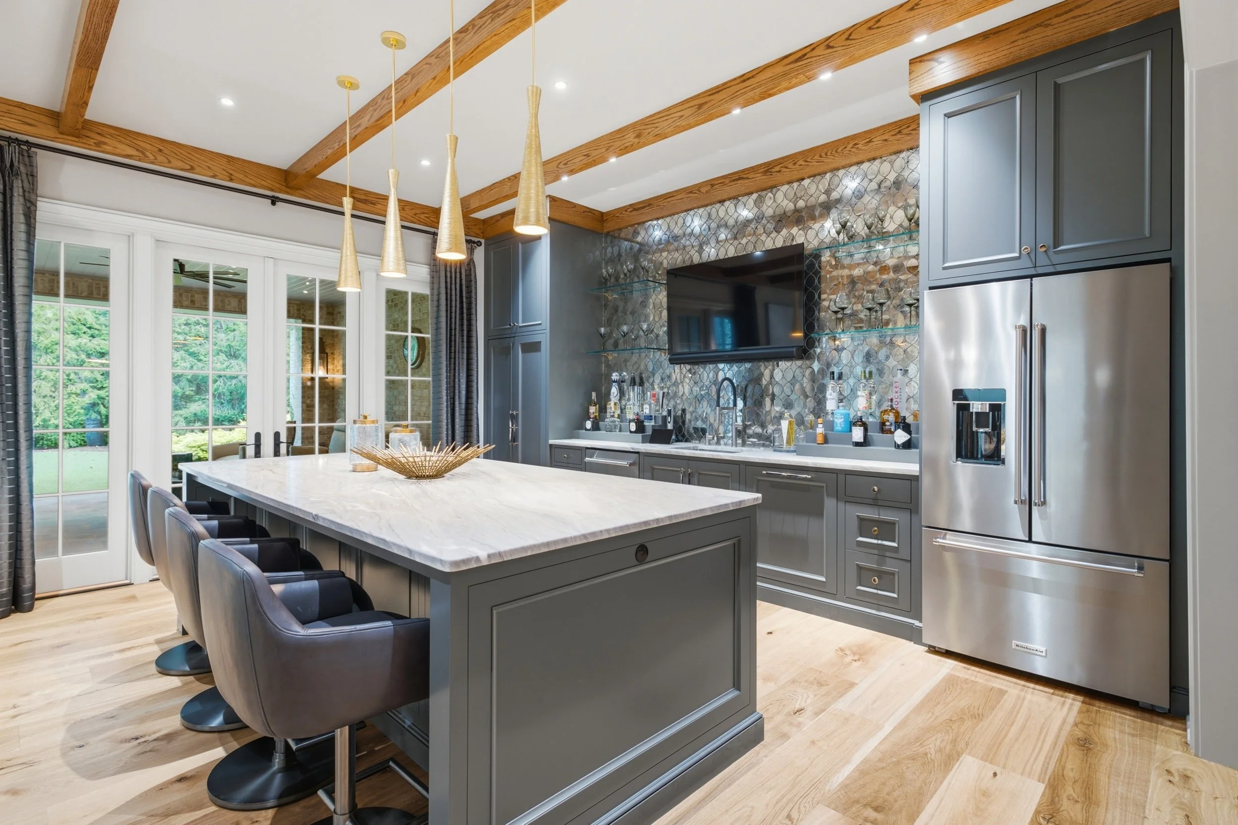 Modern kitchen with a large white marble island, gray cabinetry, pendant lights, and a view of a backyard with green trees through large glass doors.