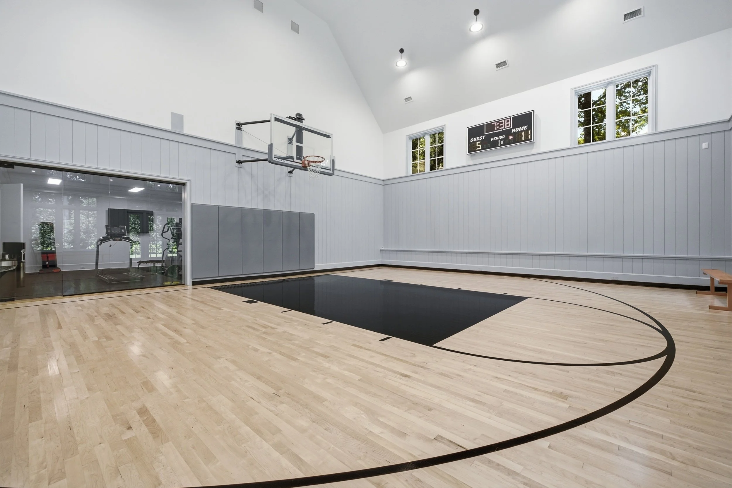 An indoor basketball court with a wooden floor, black lines, a mounted basketball hoop, and a digital scoreboard displaying 7:38. Behind a glass wall, exercise equipment and a room with a window are visible.
