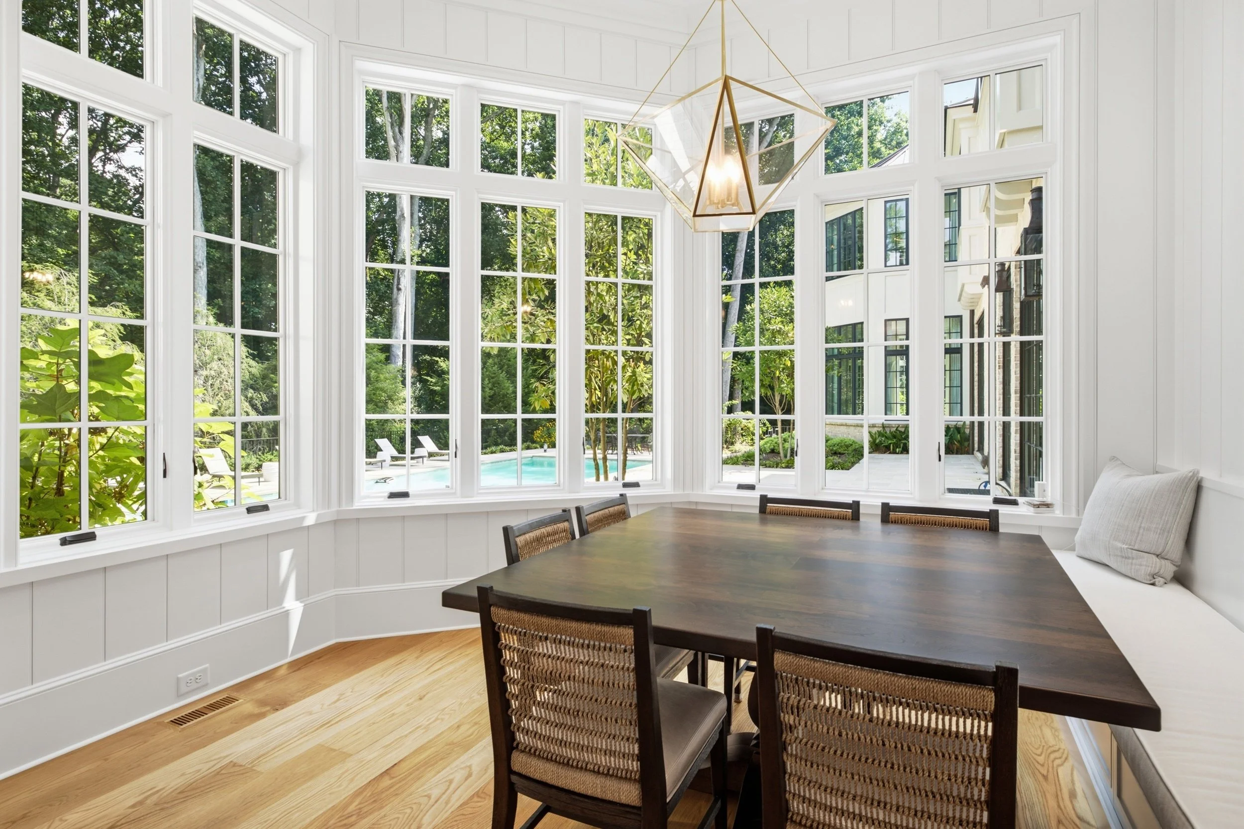 A bright dining area with large bay windows overlooking a backyard with a swimming pool. The room features a wooden dining table, wicker chairs, and a modern gold geometric pendant light fixture.