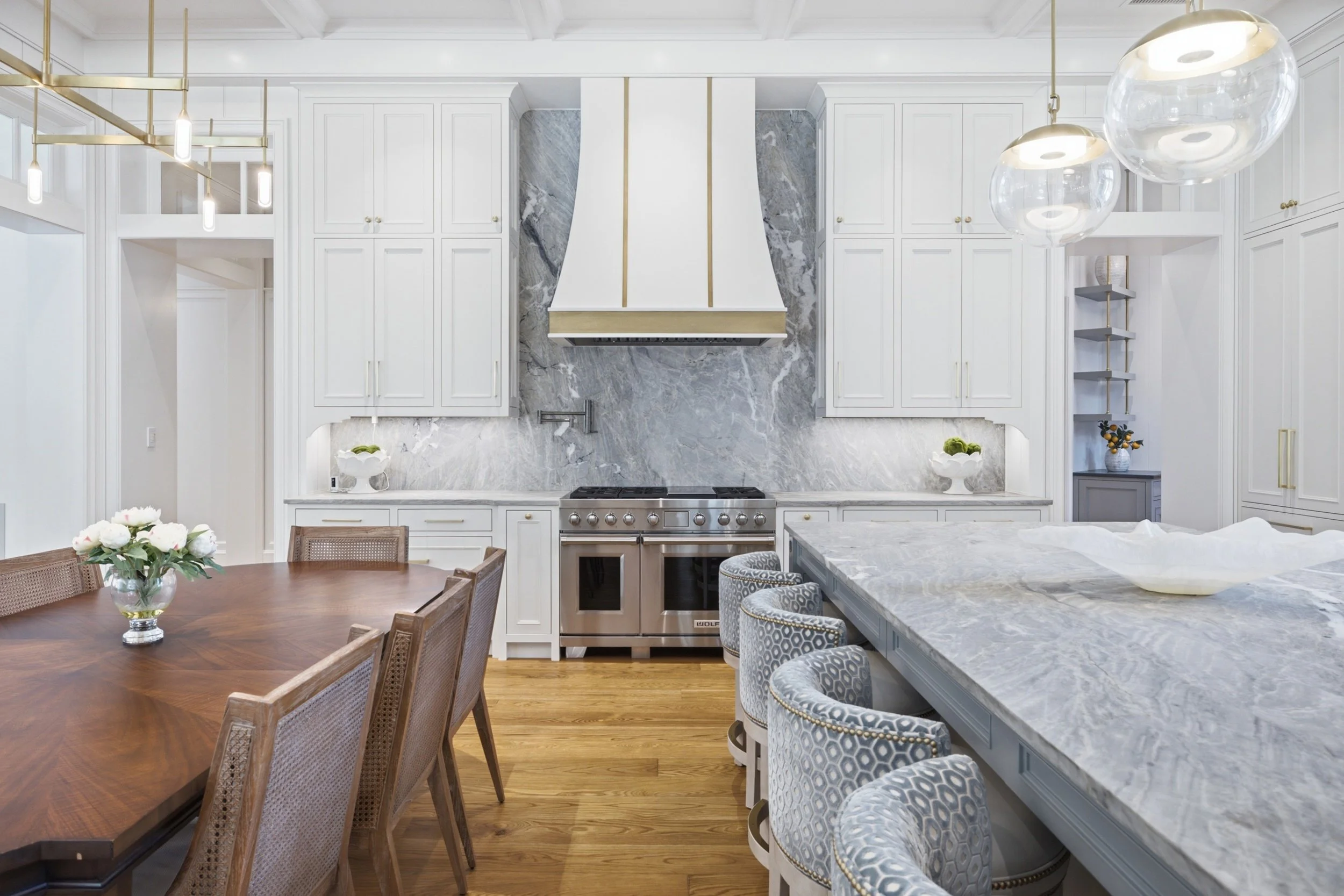 A modern kitchen with white cabinets, marble countertops, a metal stove, and gold accents. A dining table with wooden chairs and a floral centerpiece is in the foreground.