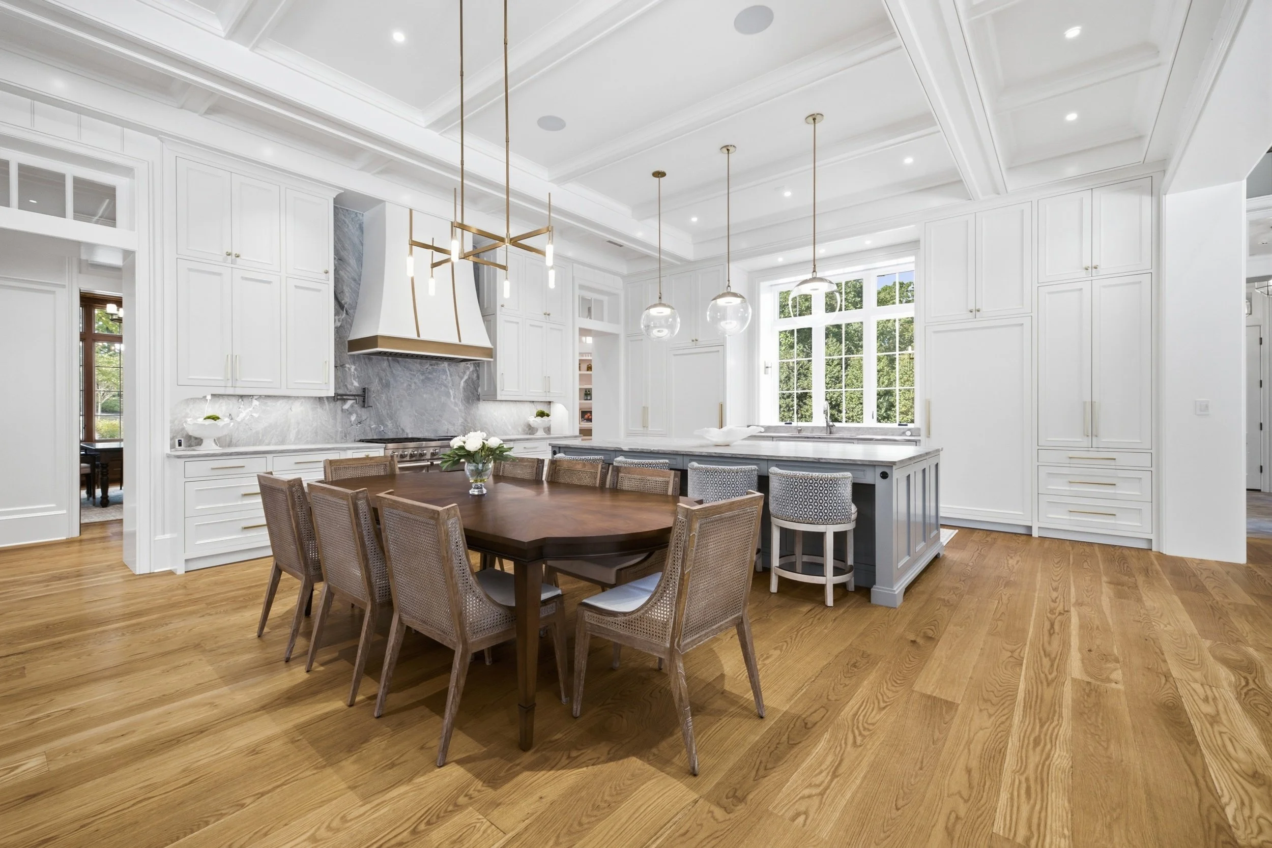 A spacious kitchen with white cabinetry, a large island with seating, wooden flooring, pendant lights, and a window overlooking a green outdoor area.