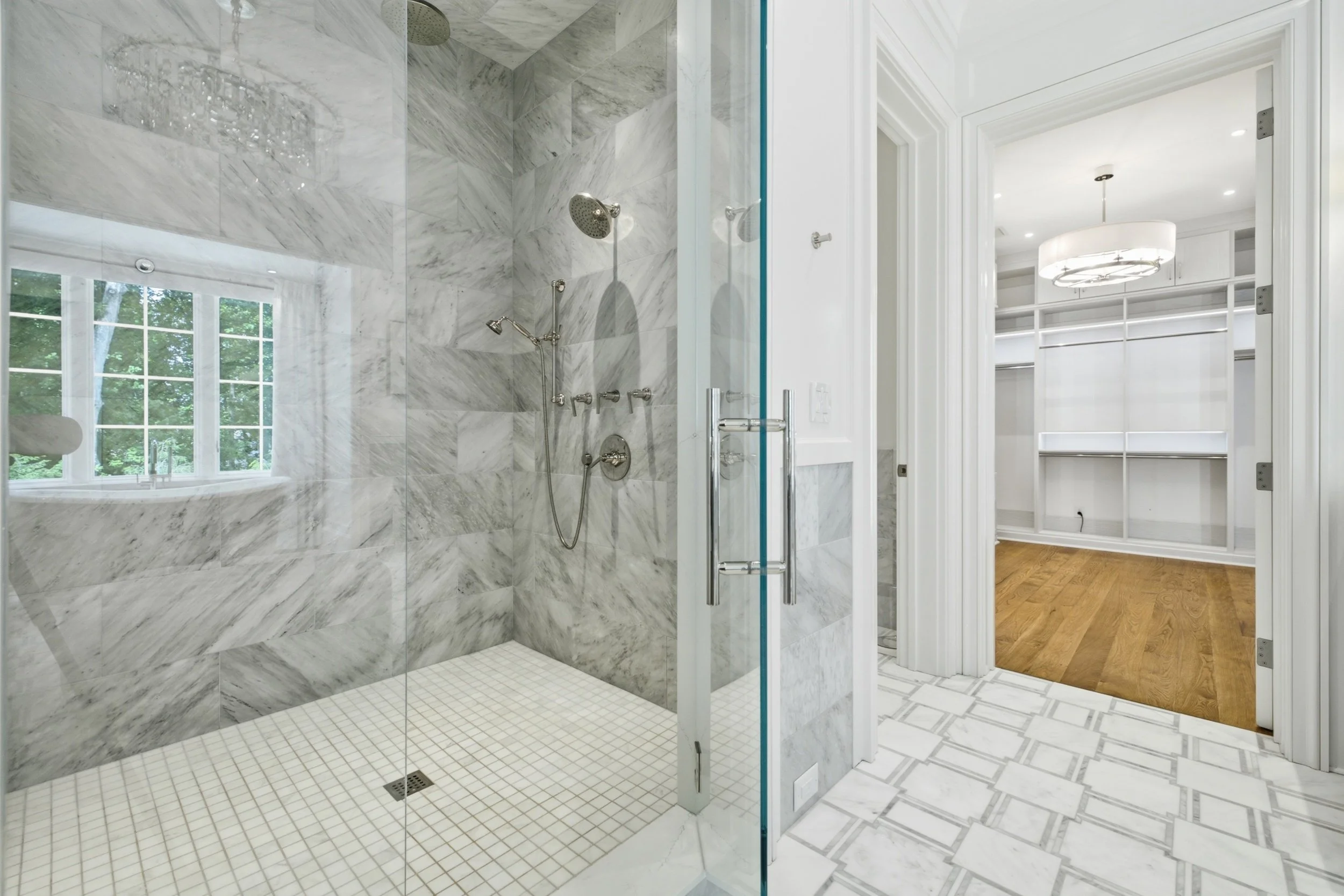 A modern bathroom with a large walk-in shower featuring marble tiles, a rain showerhead, a handheld shower, and glass doors. Inside the shower, there is a window with a view of green trees. Adjacent to the shower is a walk-in closet with white shelvi