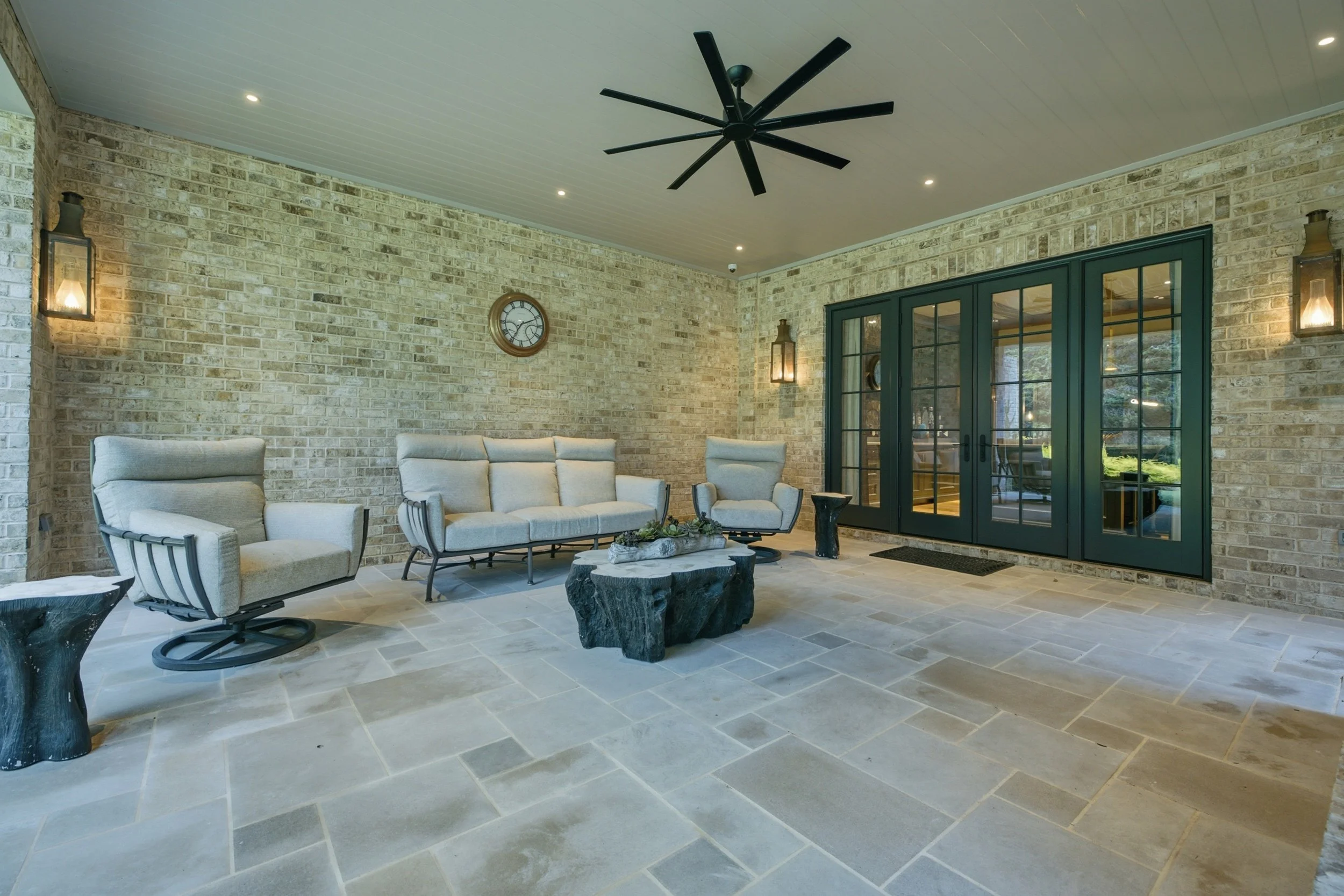 A covered outdoor patio with brick walls, a black ceiling fan, wall-mounted lantern lights, beige outdoor furniture, and sliding glass doors.