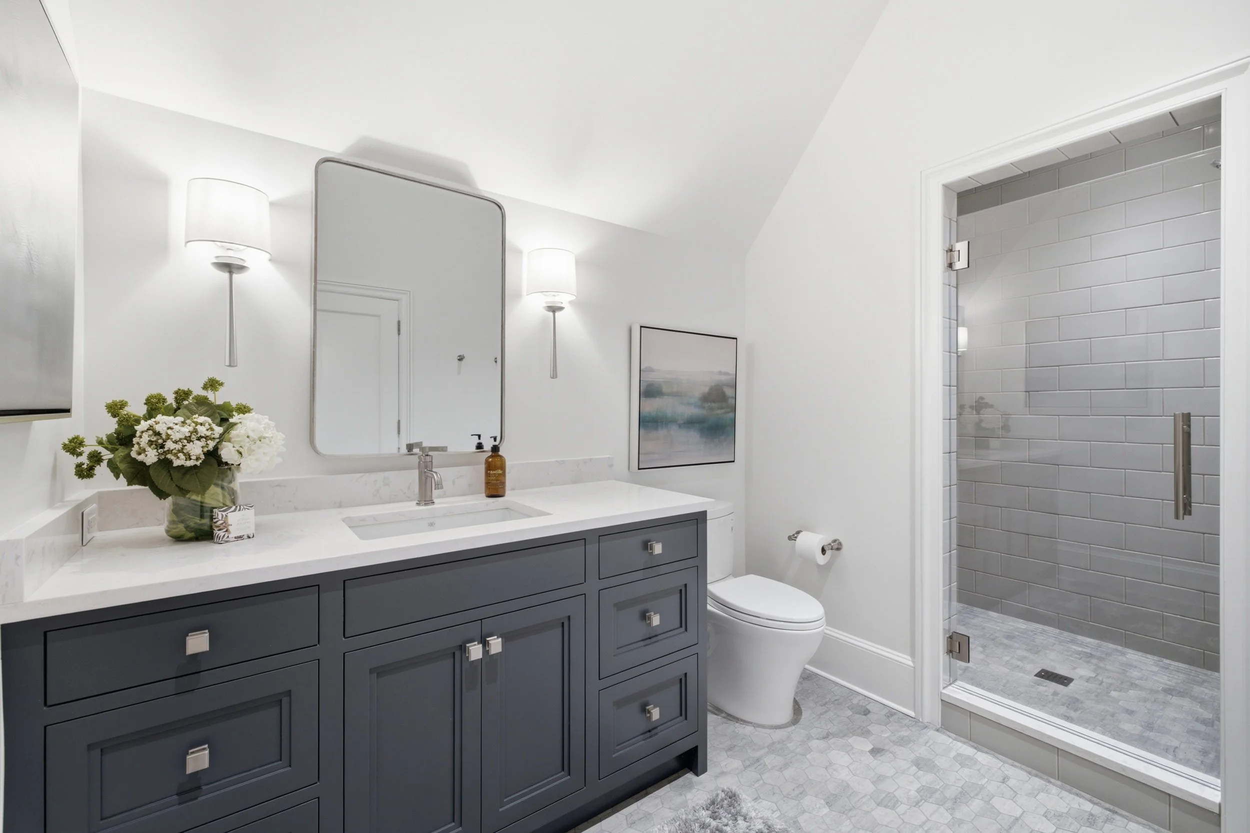 Modern bathroom with blue vanity, white marble countertop, large mirror, wall-mounted lights, flowers, soap dispenser, toilet, and a glass-enclosed shower with gray subway tiles.