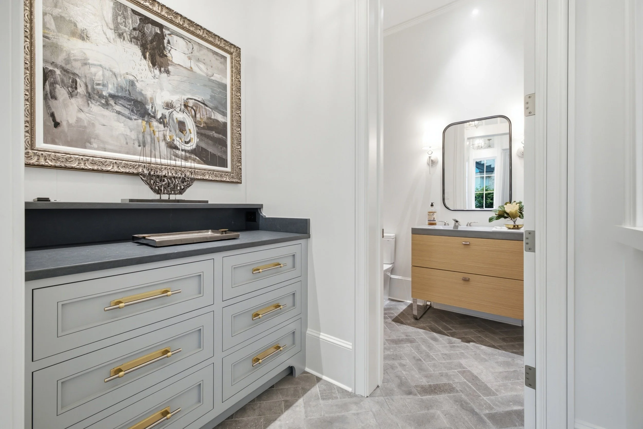A bathroom with light gray cabinetry, gold handles, a dark gray countertop, and a tray. There's a large abstract painting with a decorative frame hanging above the vanity. In the background, a mirror, vanity with a flower arrangement, and a small roo