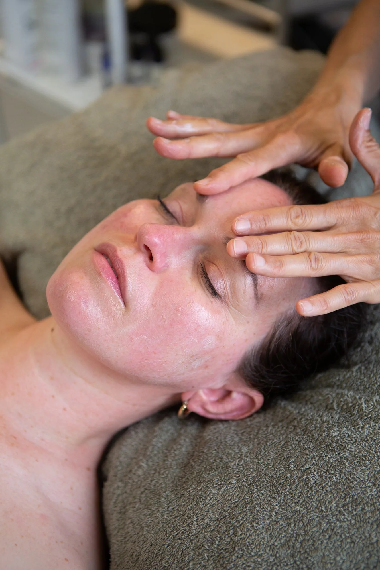 A woman lying down with her eyes closed, receiving a massage on her forehead and temples from a person using their fingers.